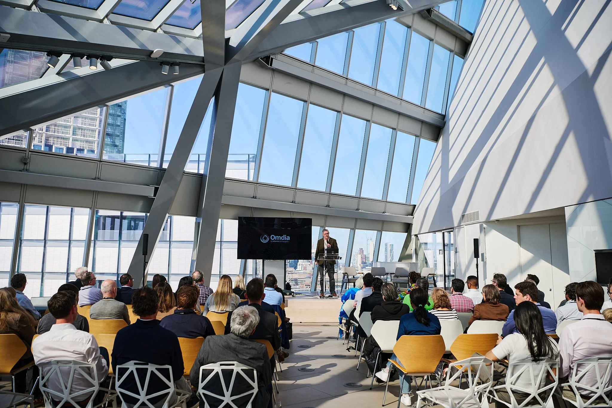 A speaker is giving a presentation at a conference inside a modern building with large glass windows and an industrial design, with an audience seated facing the stage.