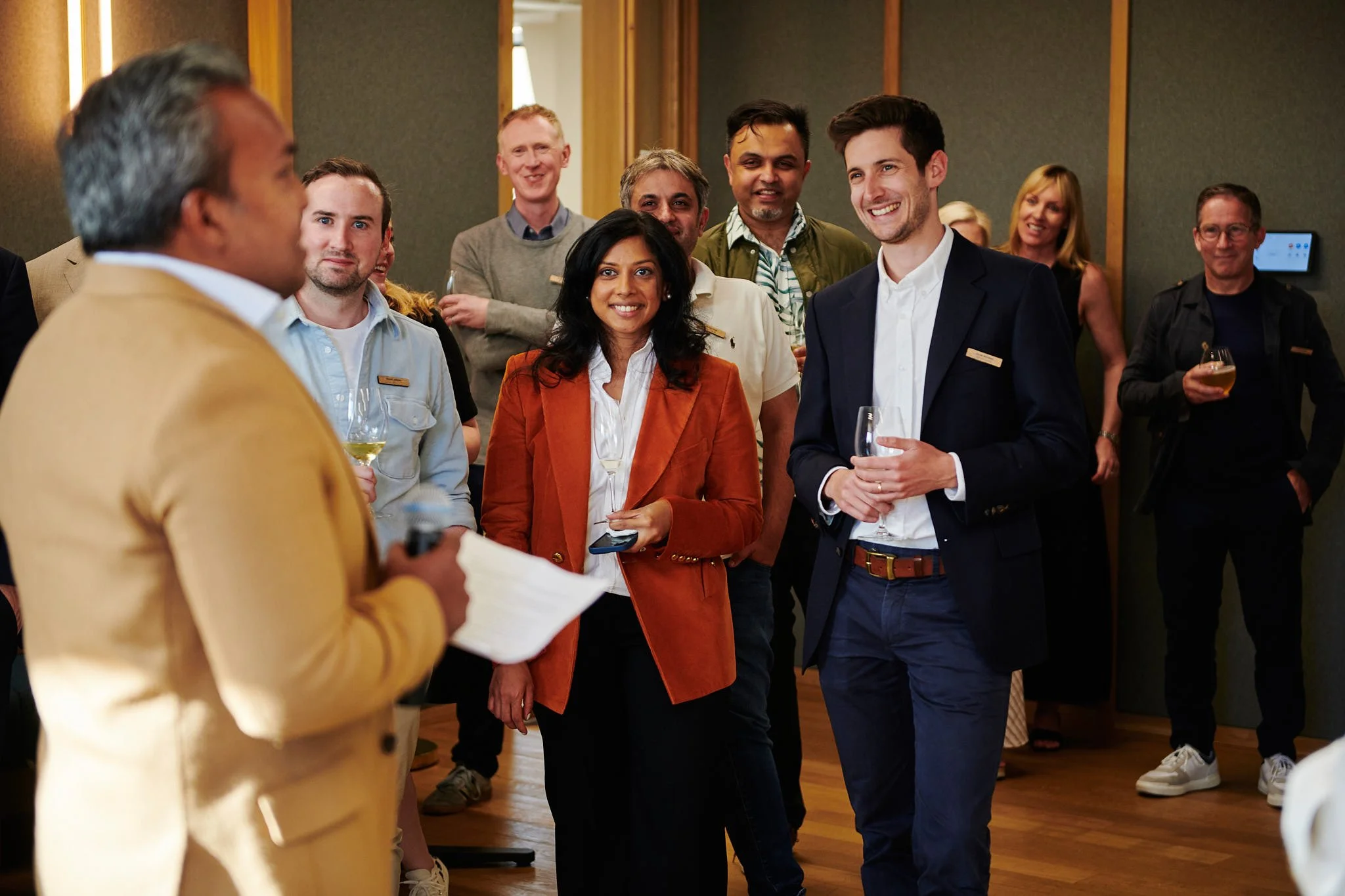 Group of professionally dressed people at a social gathering or networking event, standing indoors with drinks, engaging in conversation and smiling.