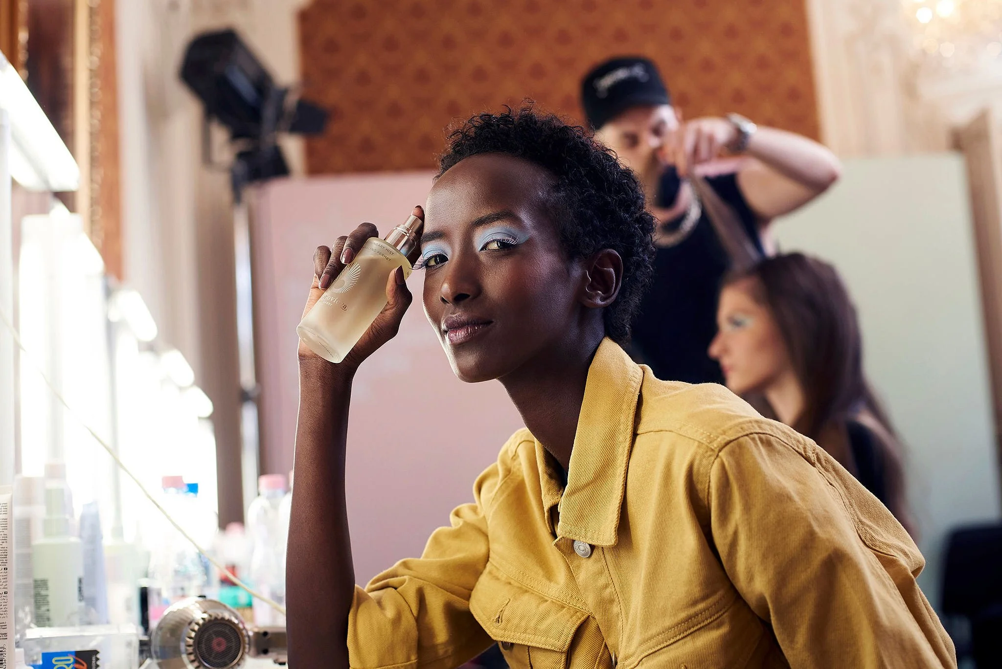 A young woman with short curly hair holding a spray bottle of perfume near her face, sitting in a room with makeup and beauty products, while two other women in the background are styling hair.