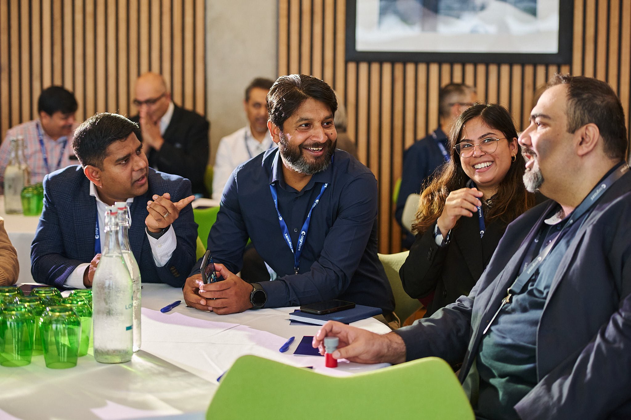 Group of diverse business professionals conversing and smiling at a conference table in a modern, wooden-paneled conference room.