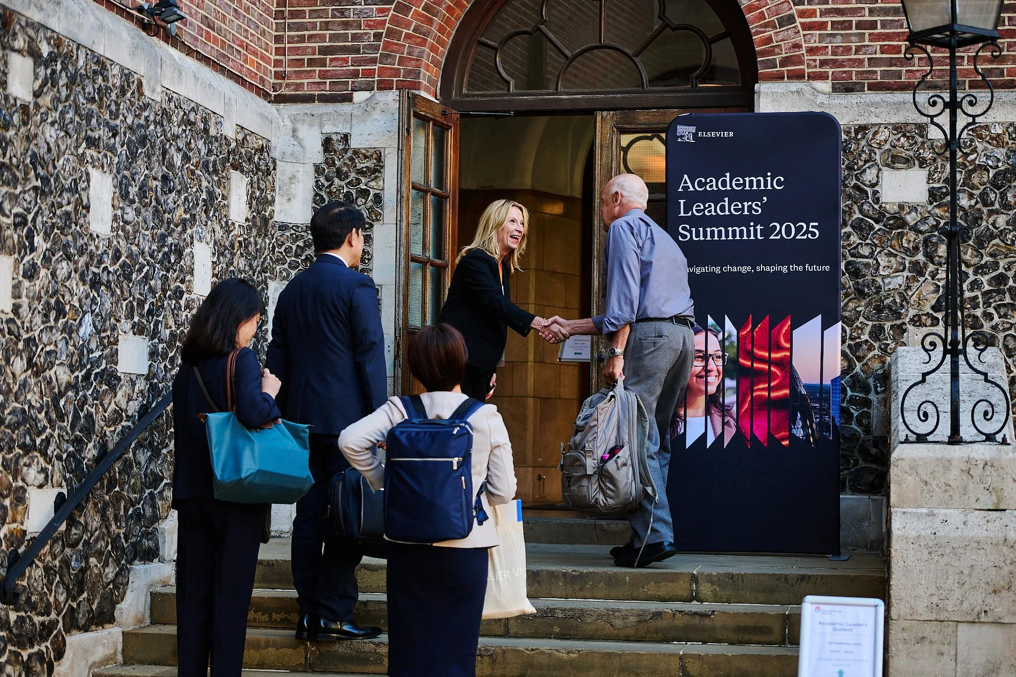 People waiting to enter the Academic Leaders' Summit 2025, with two people shaking hands at the entrance, outside a historic building.