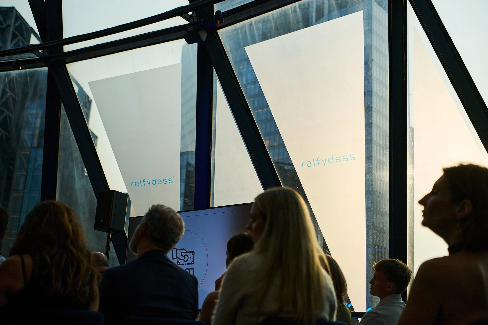 People attending a conference or event inside a modern building with large glass windows, with the word 'relfydess' displayed on the windows.