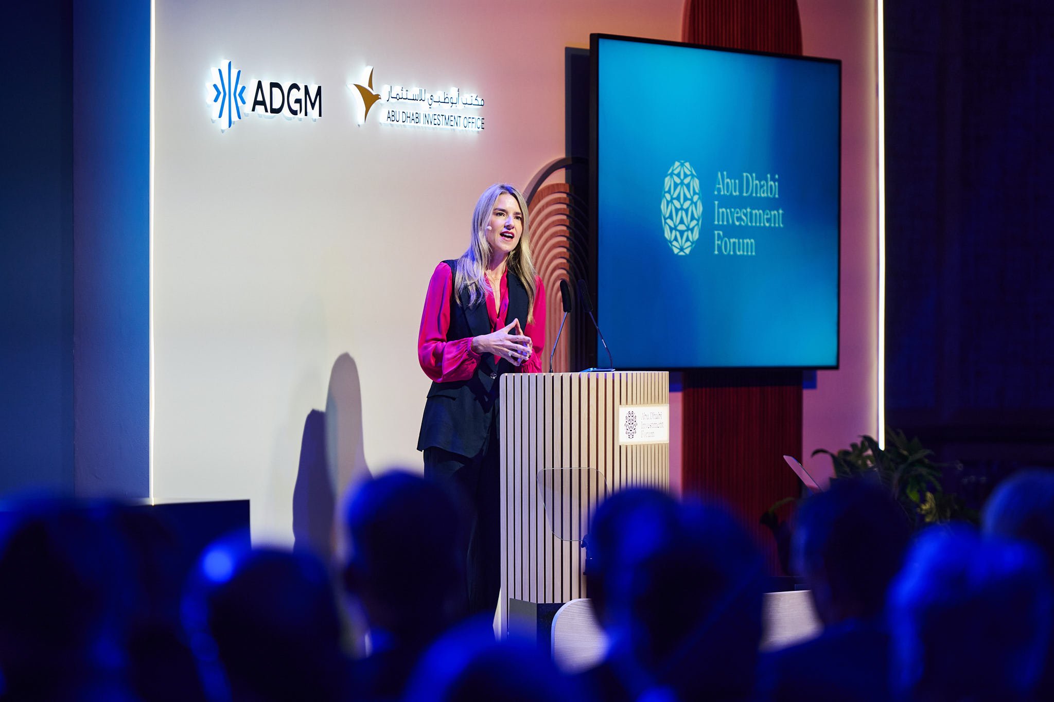 A woman speaks at a podium during a presentation at the Abu Dhabi Investment Forum, with the event's logo on a screen and the Abu Dhabi Investment Office and ADGM logos on the wall behind her.