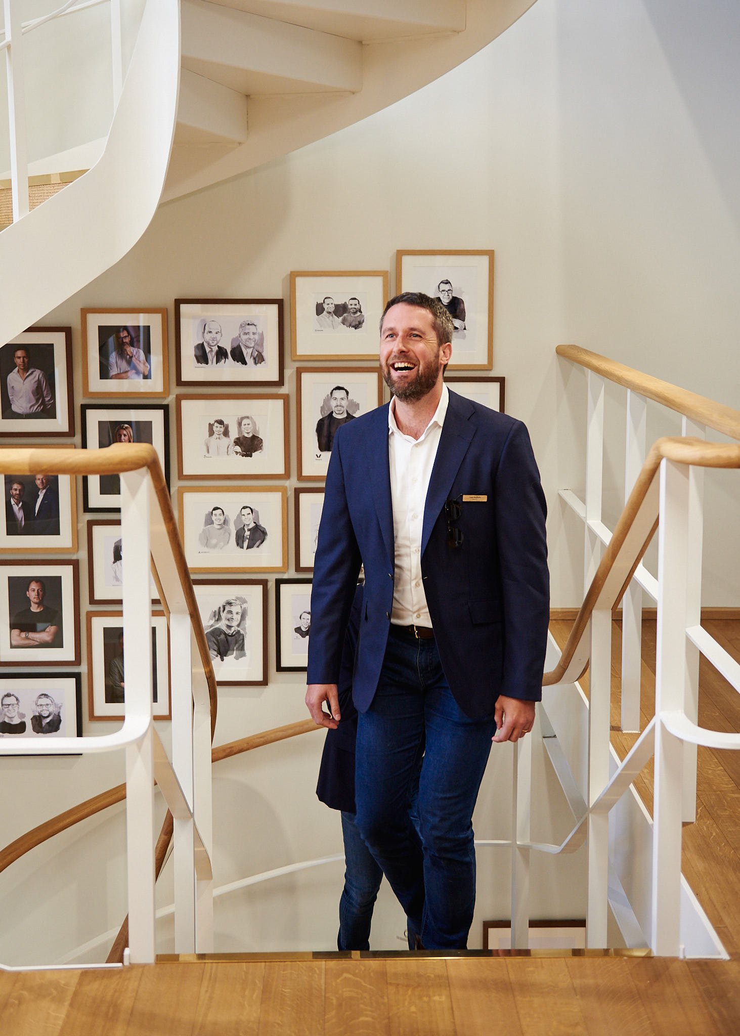 A man in a navy blue blazer and white shirt standing on a staircase, smiling, with framed photos on the wall behind him.