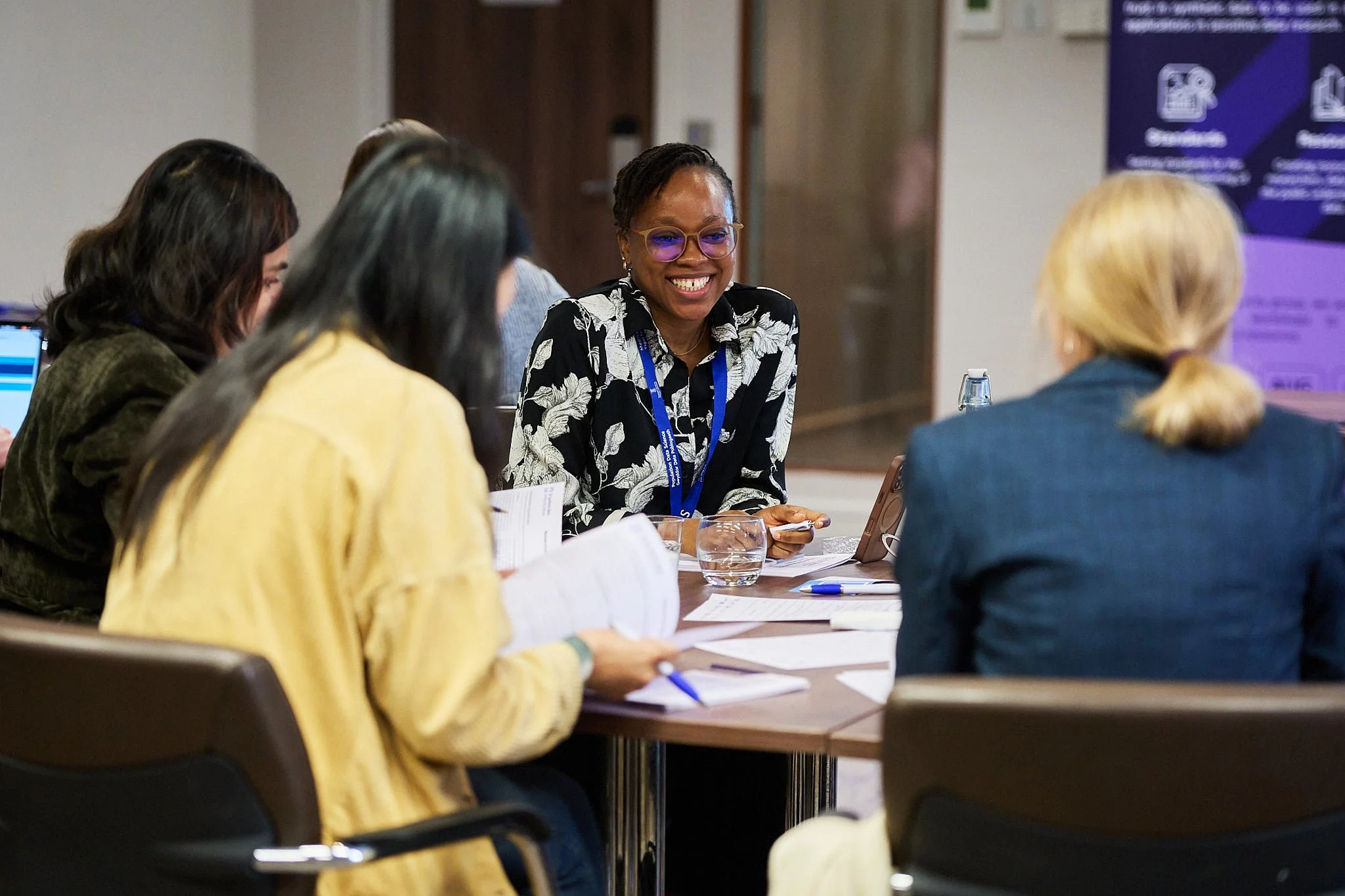 Group of women in a meeting room, engaged in discussion, with one woman smiling and wearing glasses and a floral shirt.