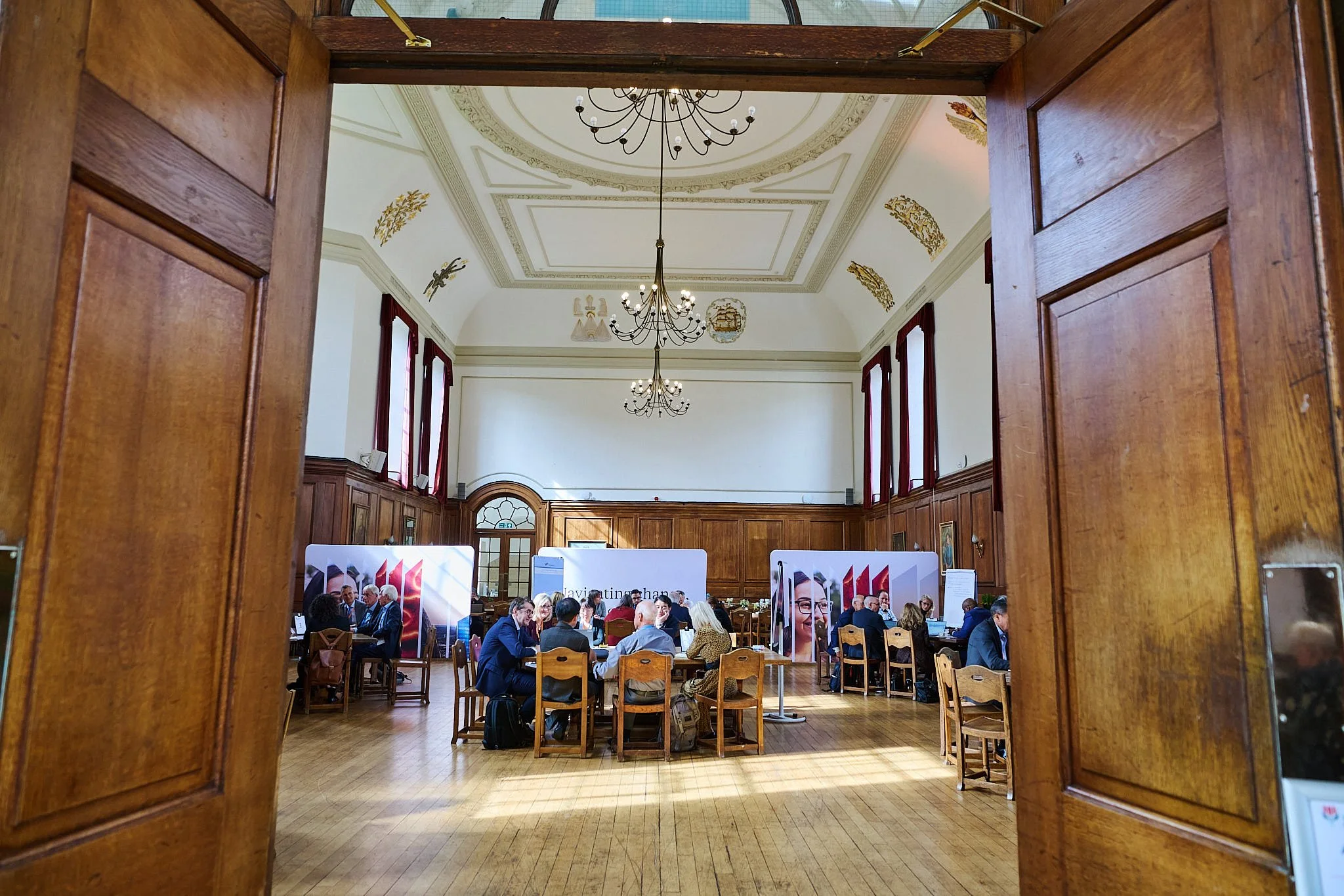 A spacious conference room viewed through open wooden doors. The room has a high, decorated ceiling with chandeliers and ornate gold decorations. People are seated at round tables, engaging in discussions, with exhibition stands along the walls displ