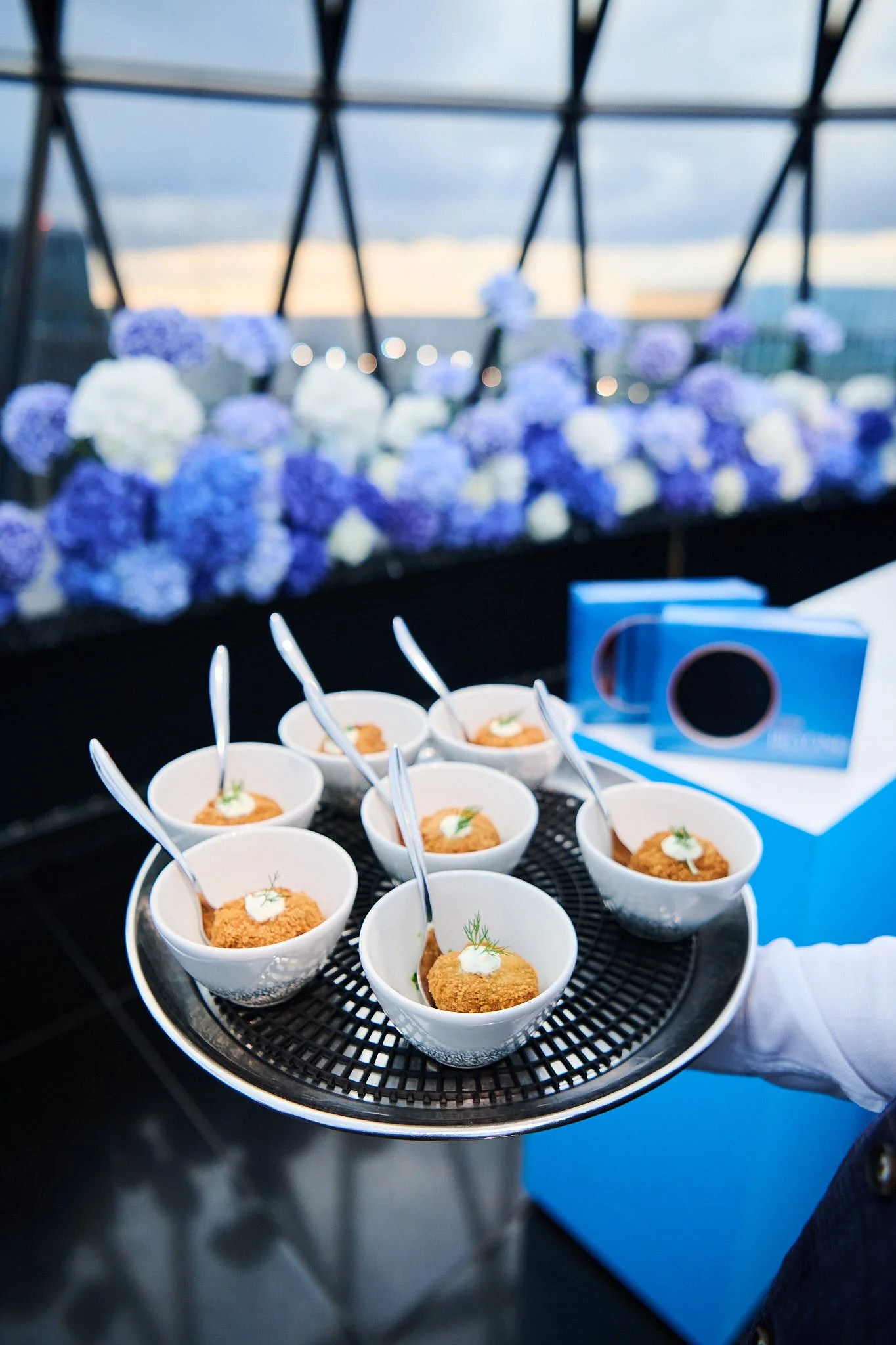 Tray with six small bowls of appetizers, with spoons, on a rooftop with purple and white flowers in the background.