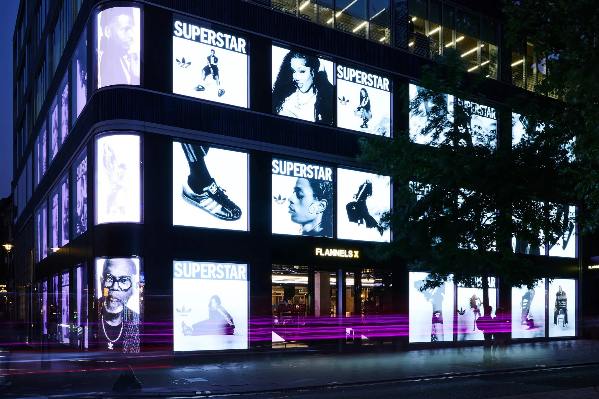 Night view of a modern building with illuminated advertising screens displaying images of various people, including a man and woman, with the word 'SUPERSTAR' on several signs. The building has multiple platforms of glass with tracks of pink and purp