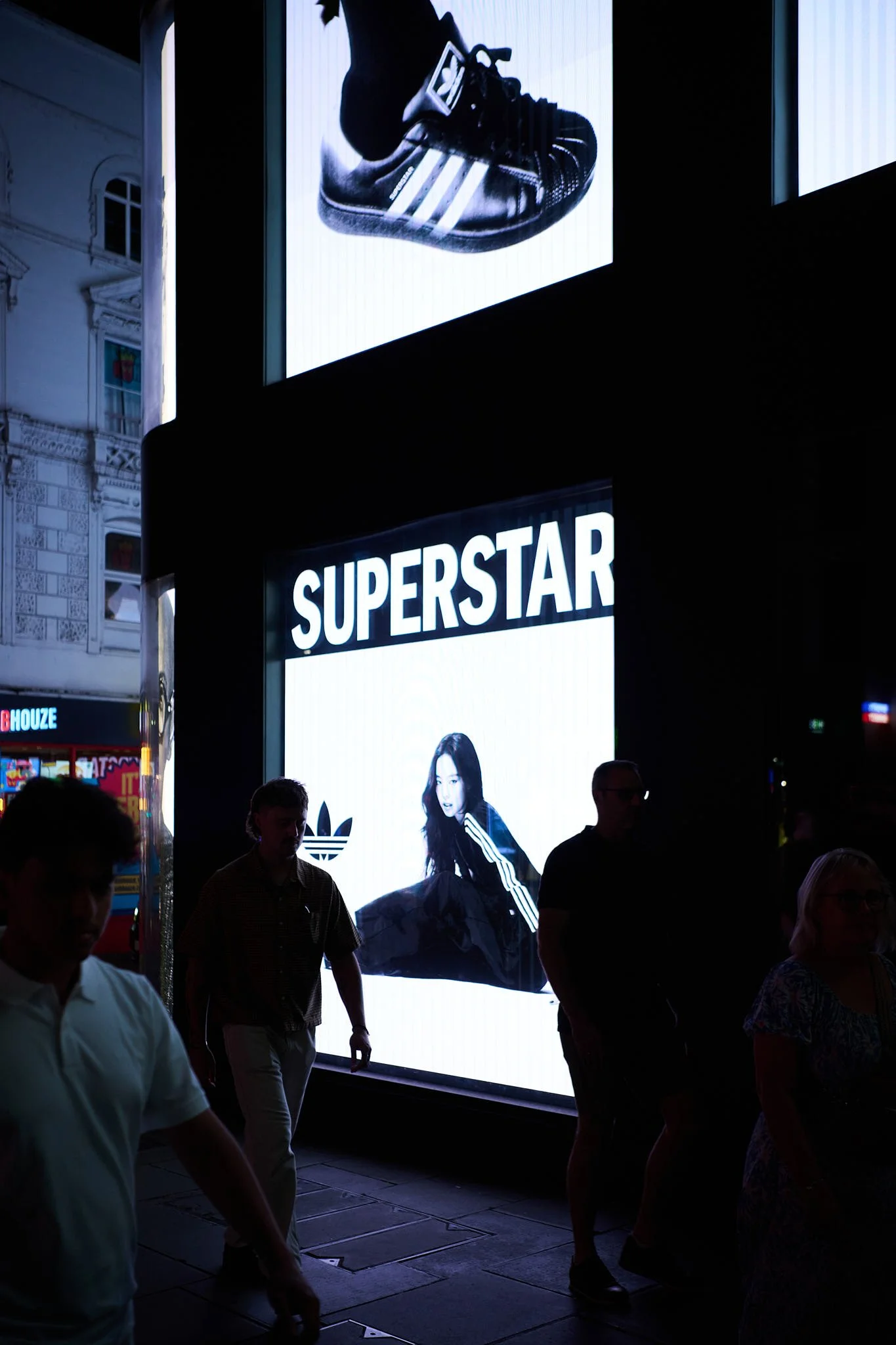 Bright billboard with the word "SUPERSTAR" and a woman in athletic attire, along with a shoe advertisement, in an urban setting at night with silhouetted pedestrians.