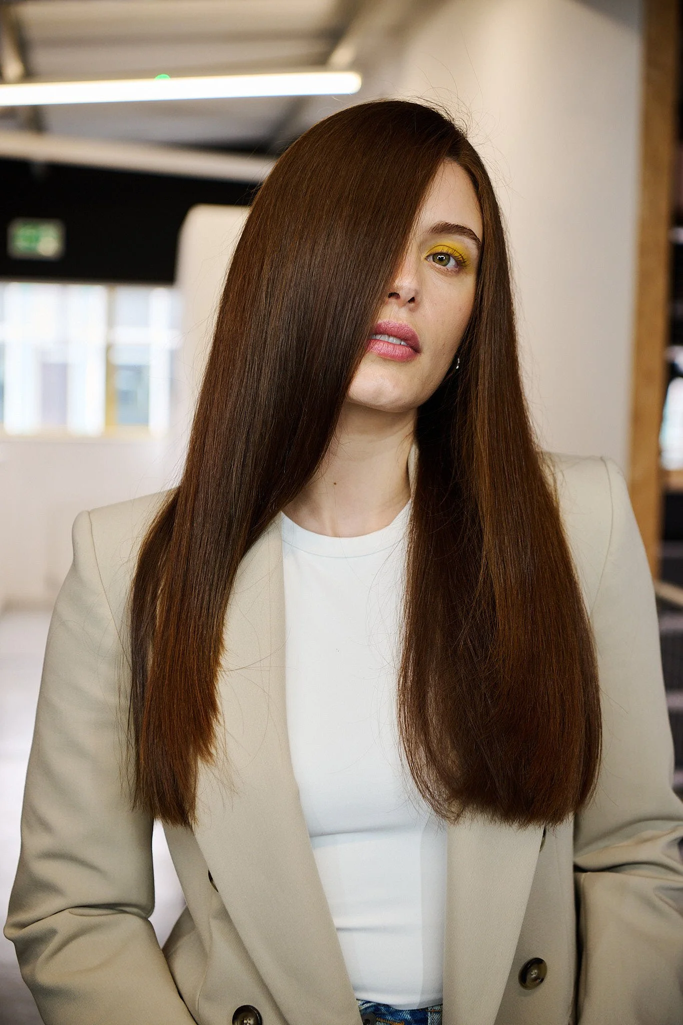 A woman with long, straight brown hair partially covering her face, wearing a cream blazer and white top, in an office setting.