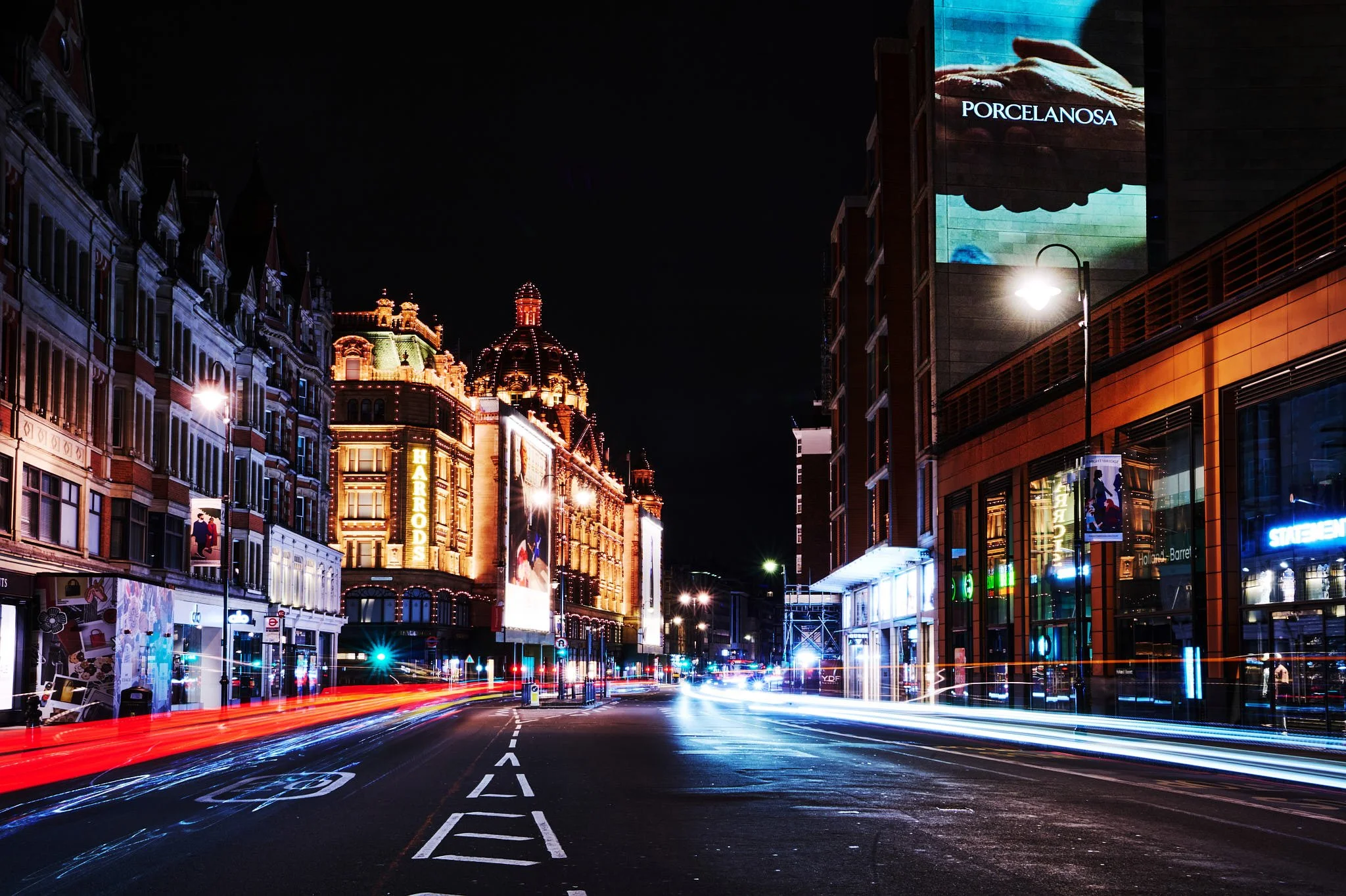Nighttime city street with illuminated historic buildings on the left and modern storefronts on the right, red and white light trails from passing vehicles, and large digital billboards on the buildings.