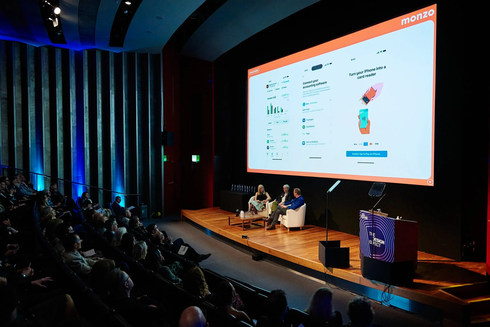 A conference room with a stage and three panelists seated on white chairs, presenting to a large audience. A big screen behind them displays a slide about Monzo's mobile app features.