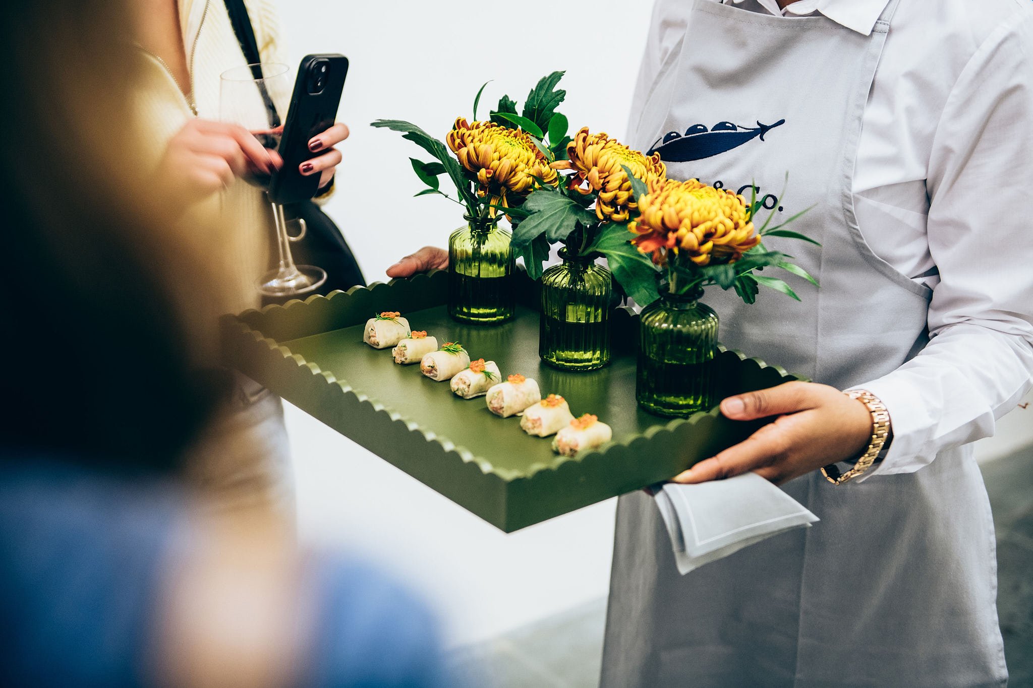 A server in a white apron holding a tray with yellow flowers in green vases and small appetizers, while a woman in a beige sweater holding a wine glass and phone stands nearby.
