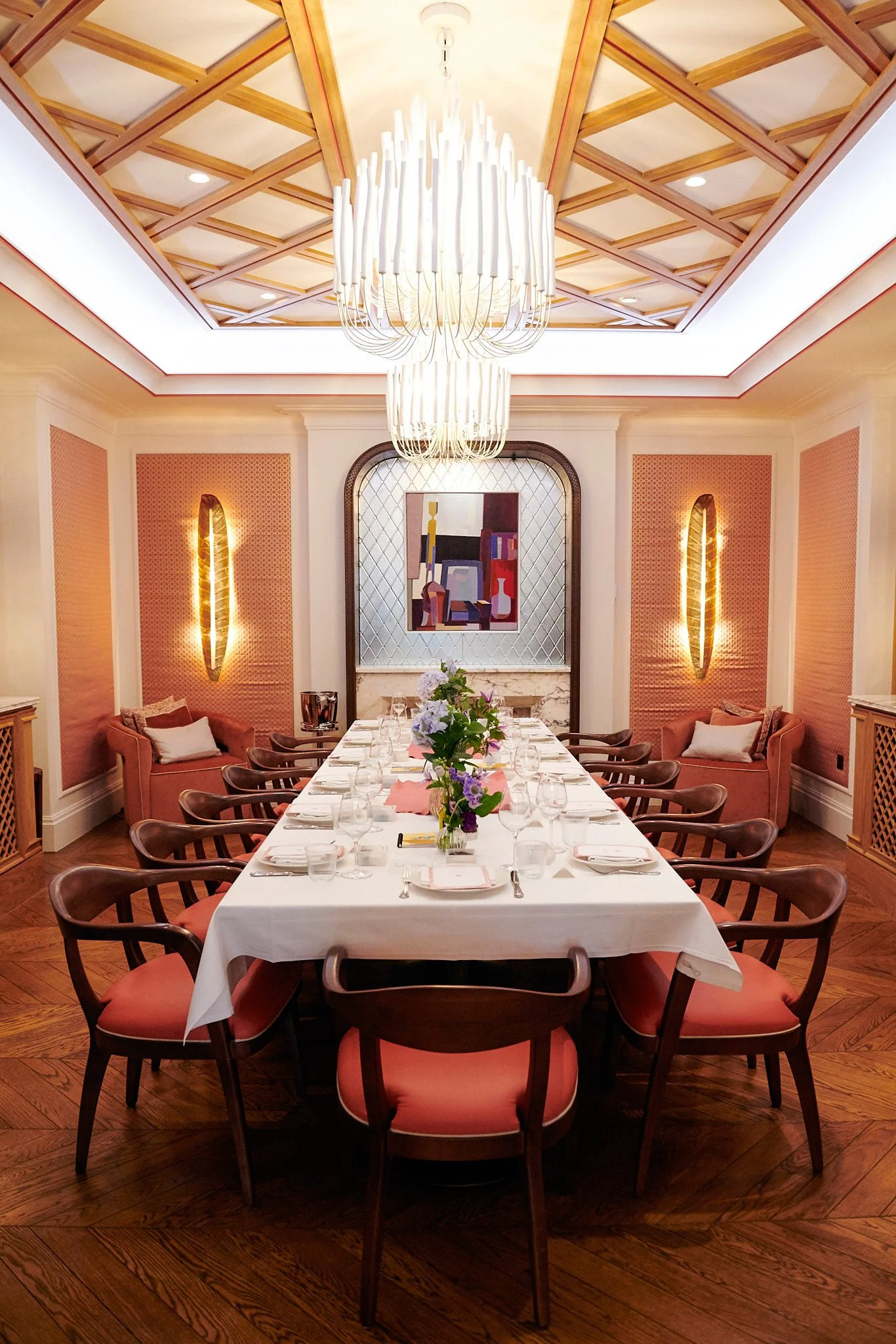 Elegant dining room with a long table set for a meal, surrounded by wooden chairs with pink cushions. The room has warm lighting, decorative wall sconces, and modern chandelier lighting fixtures hanging from a wooden coffered ceiling. There are two p