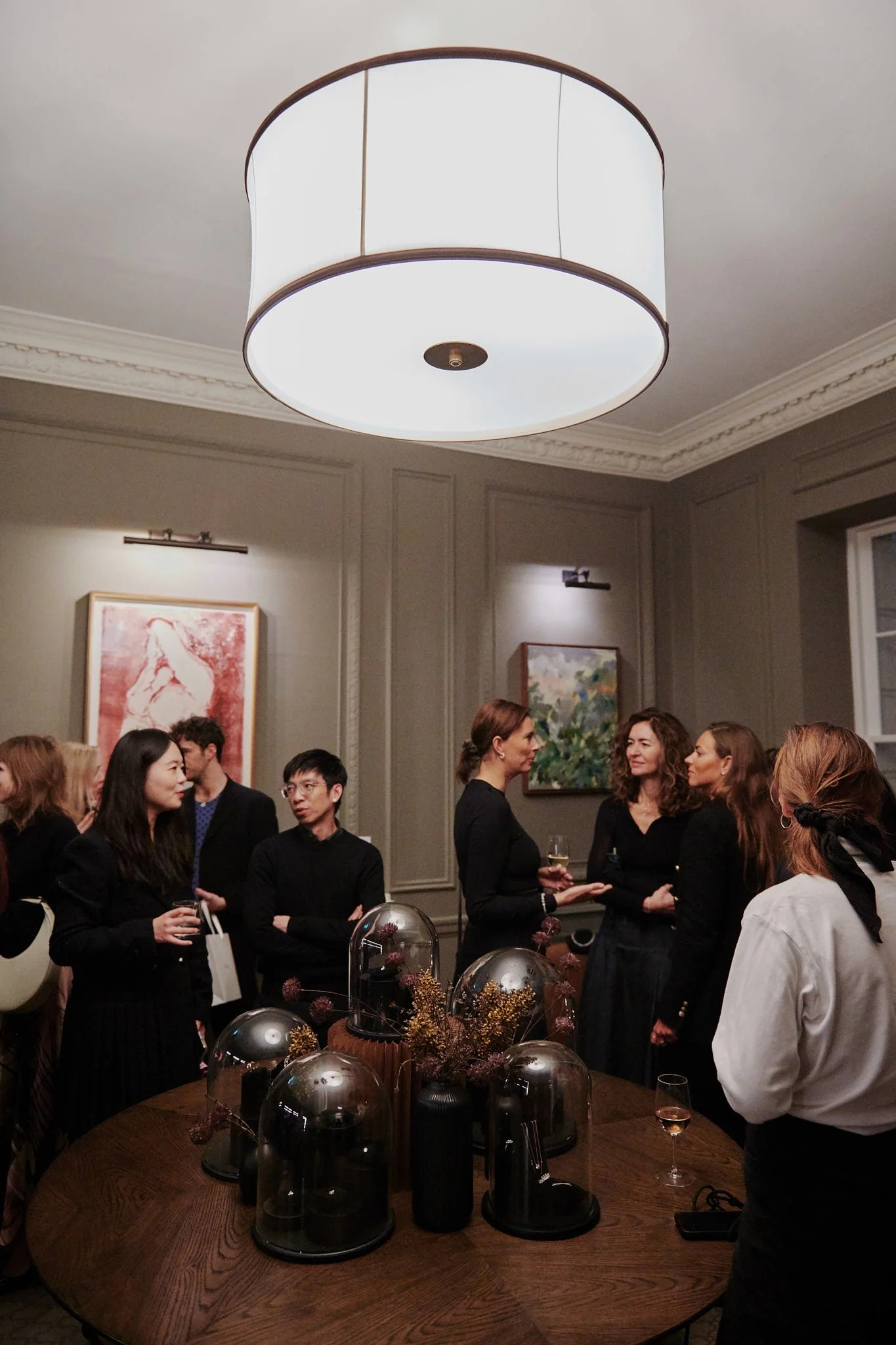 People gathered in a living room, engaging in conversation, with art on the walls, a large ceiling light, and a wooden table with decorative glass cloches and floral arrangements.