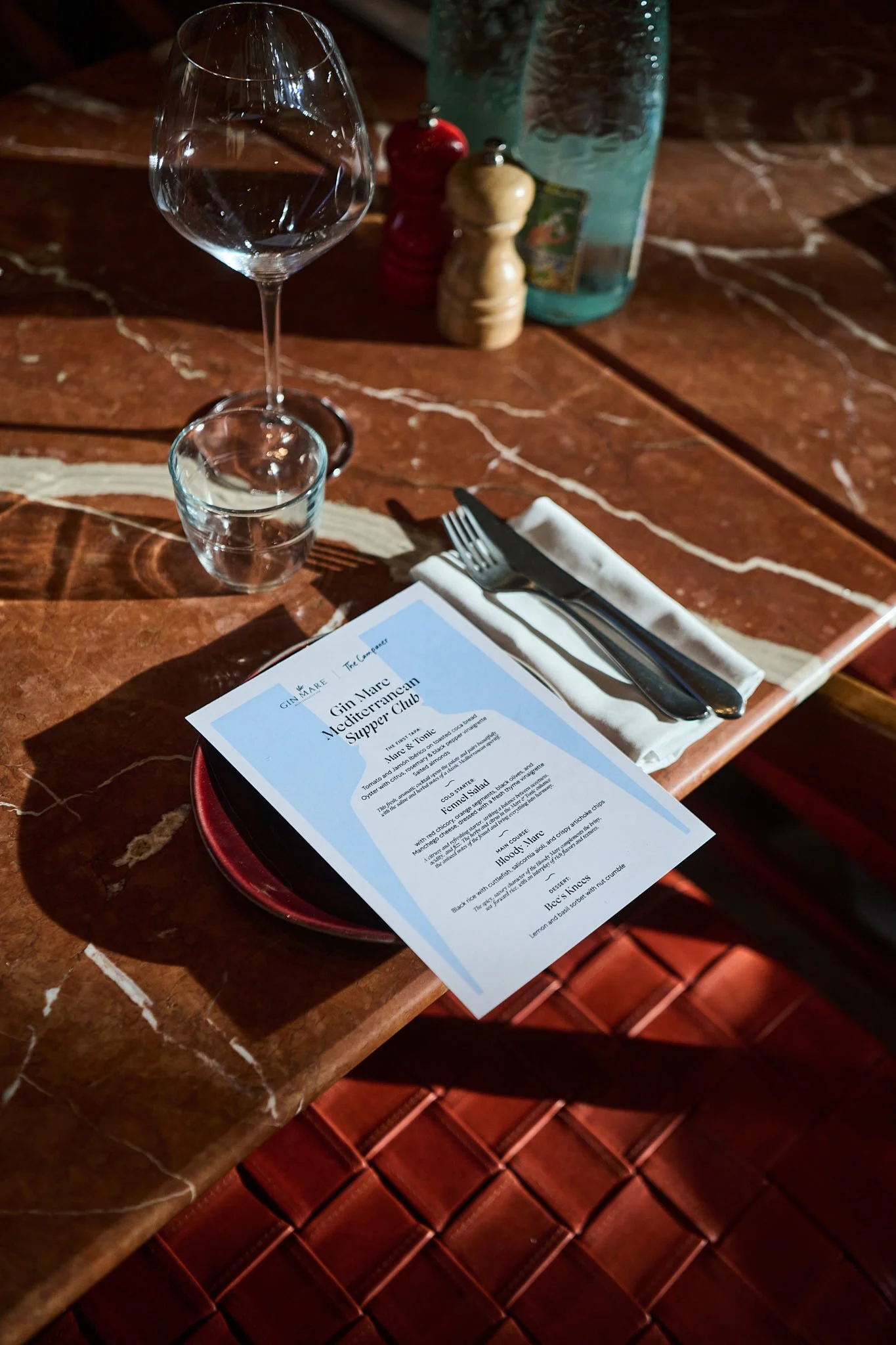 A restaurant table setting with a wine glass, a shot glass, a fork, a knife on a white napkin, and a menu titled 'Gin Tart Mediterranean Super Drink,' on a brown marble tabletop with a water bottle, salt and pepper shakers in the background.