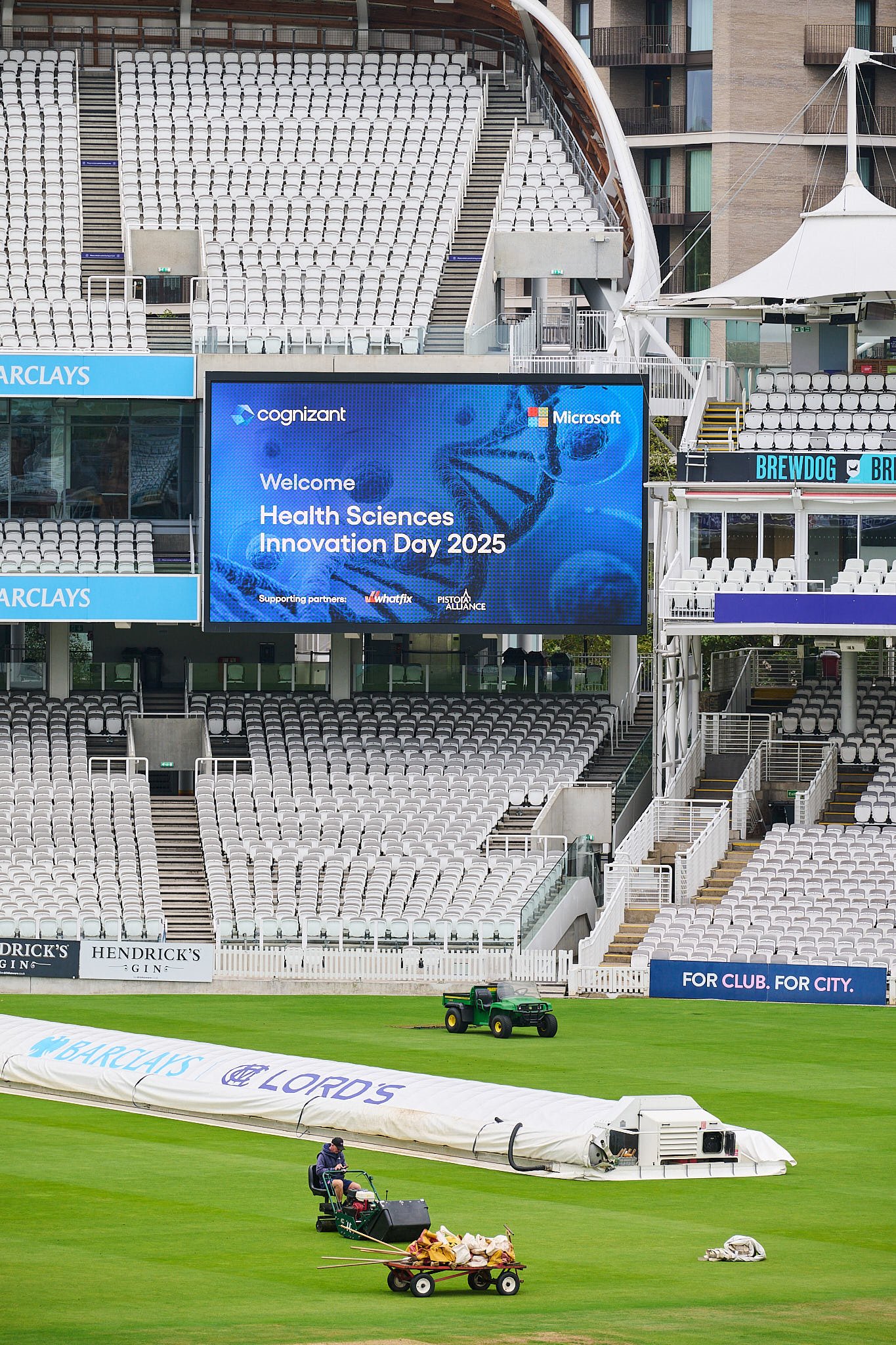 Empty stadium seating at the cricket ground with a large digital screen displaying the message for 'Health Sciences Innovation Day 2025'. The stadium features banners for Barclays and Hendrick's Gin, and a field maintenance vehicle on the grass.