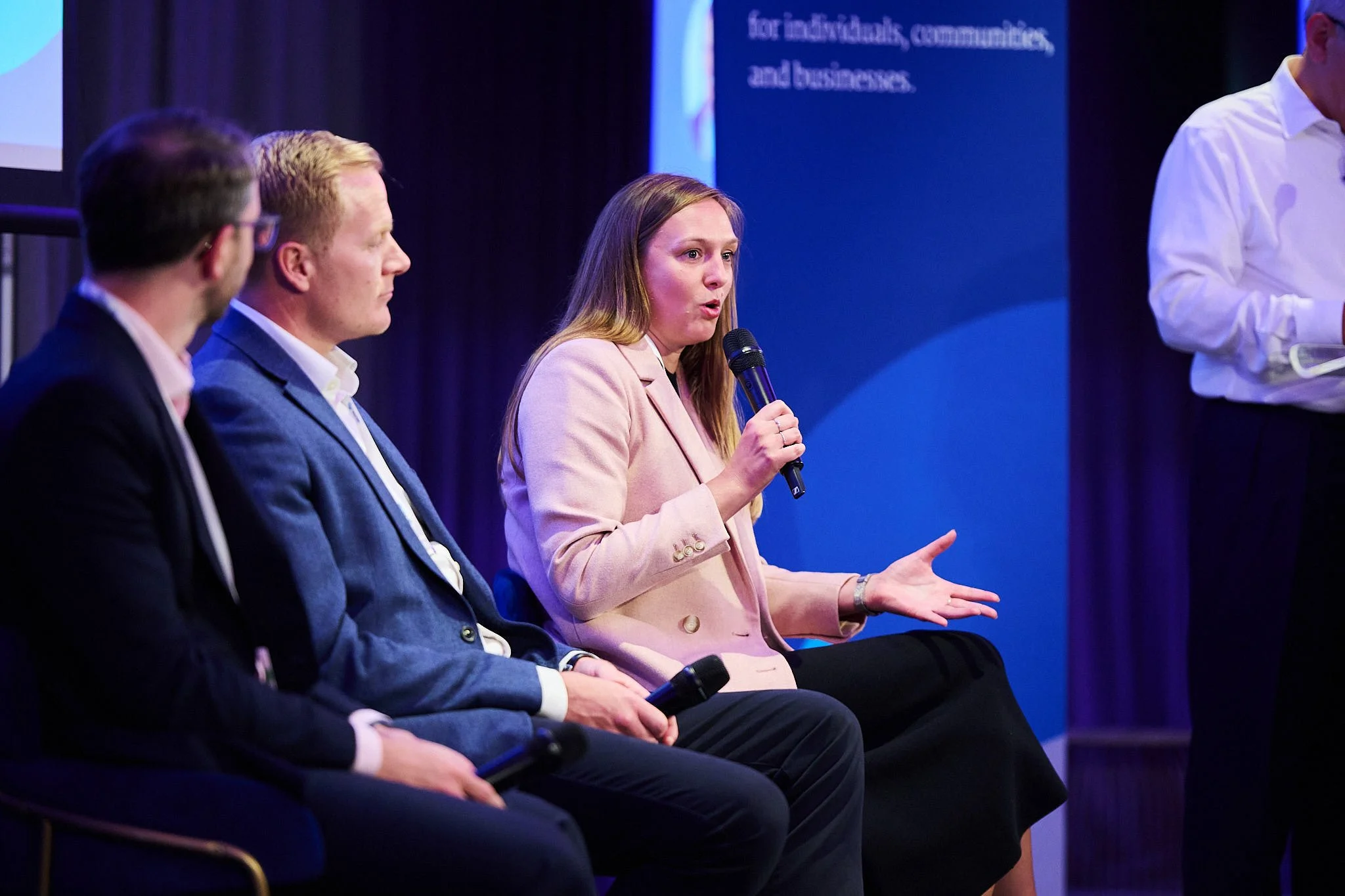 A woman in a pink blazer speaking into a microphone during a panel discussion, seated next to two men in suits. Another person stands nearby holding a device, possibly a tablet, in a professional setting with a blue backdrop.