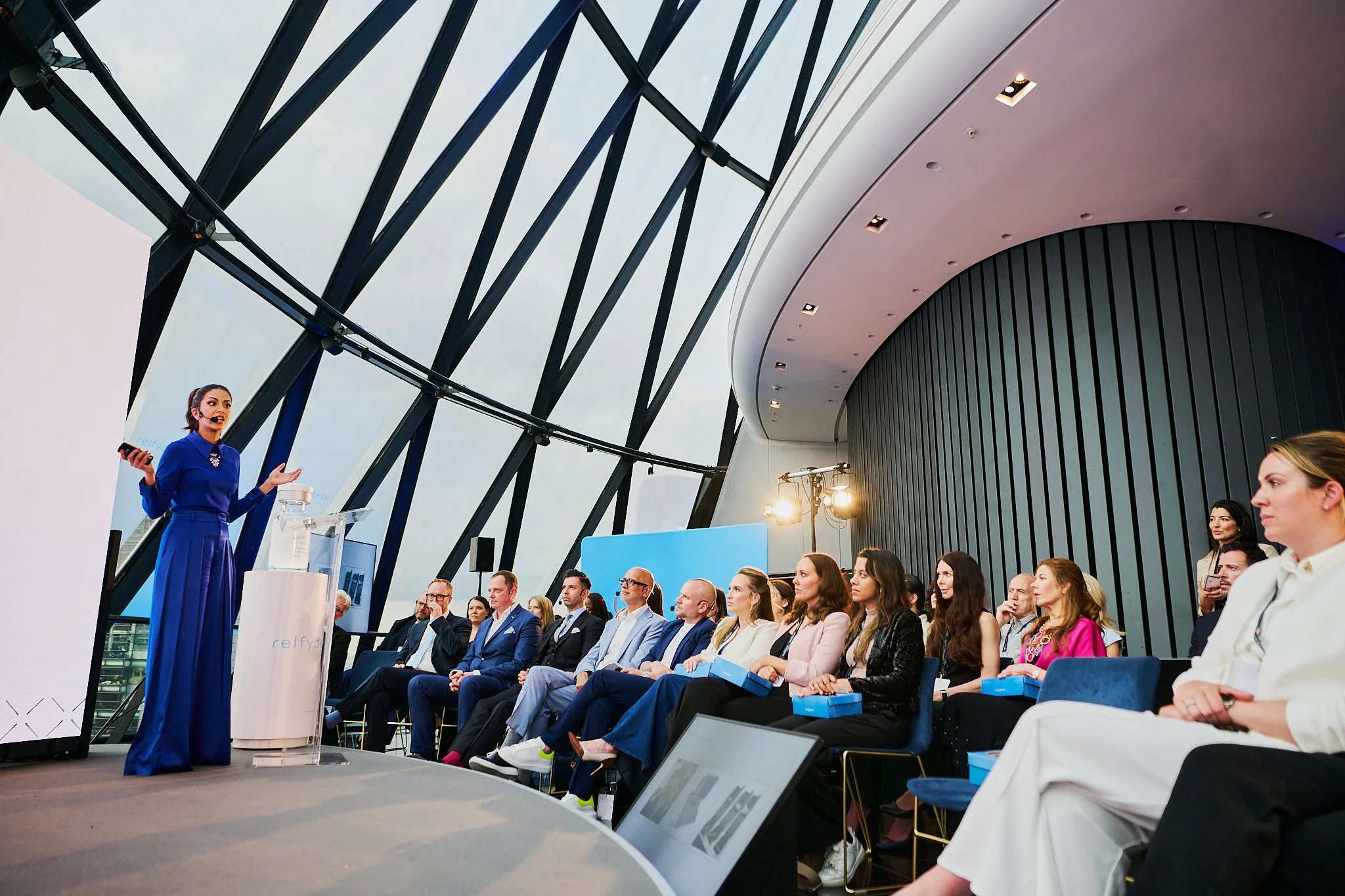 A woman in a blue dress giving a presentation to an audience seated in a modern conference room with a curved glass ceiling and dark vertical wall panels.