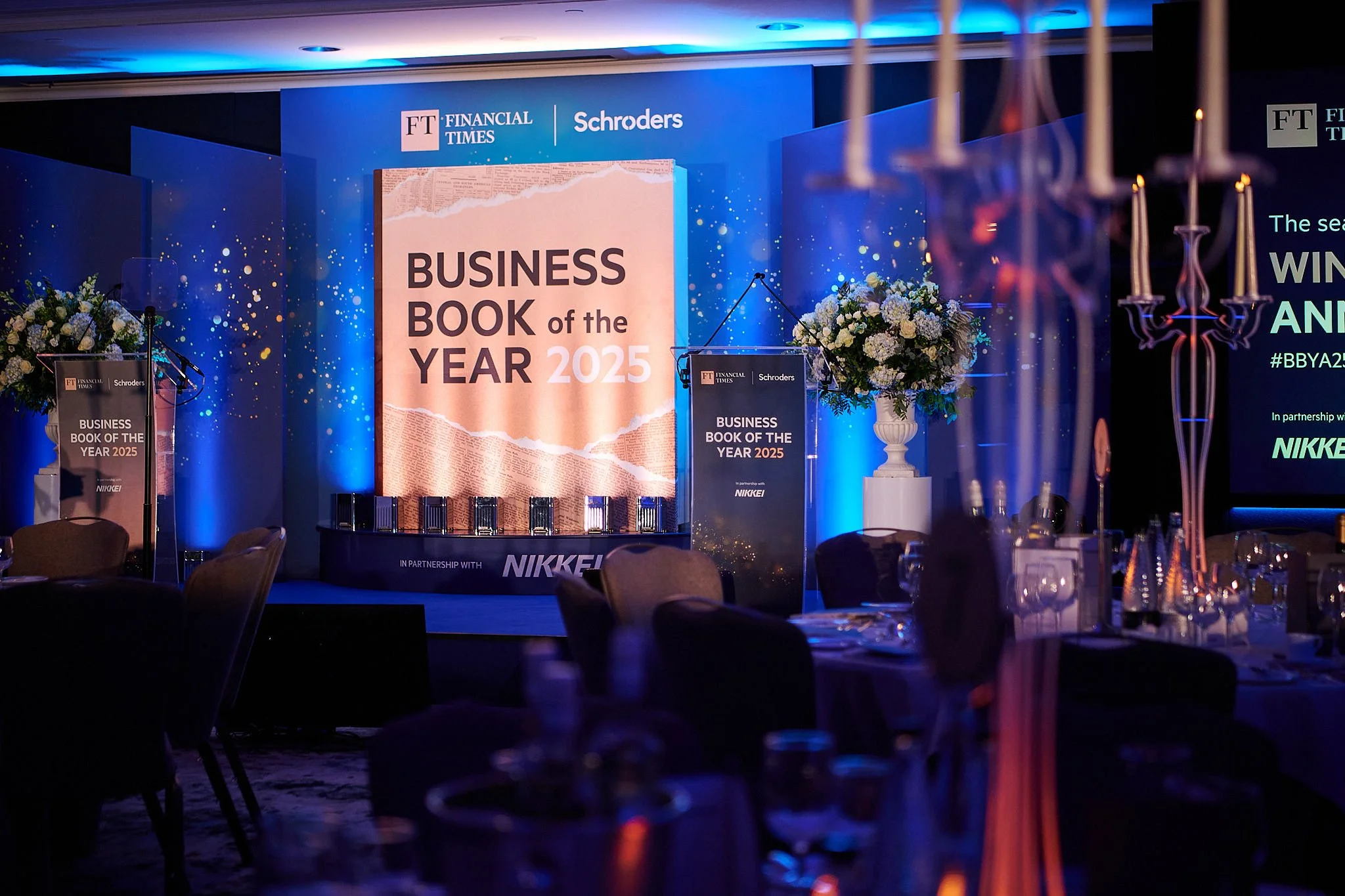Stage setup for the Business Book of the Year 2025 event with a large screen, floral arrangements, and a podium, illuminated with blue lighting.