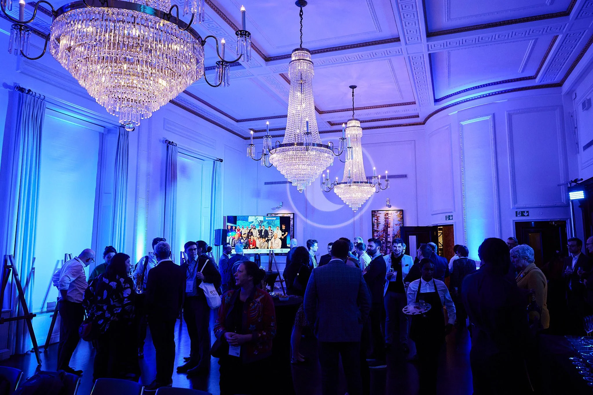 People attending a formal event in a large, elegant room illuminated with blue lighting, with three large chandeliers hanging from the ceiling, and a large screen displaying a group photo.