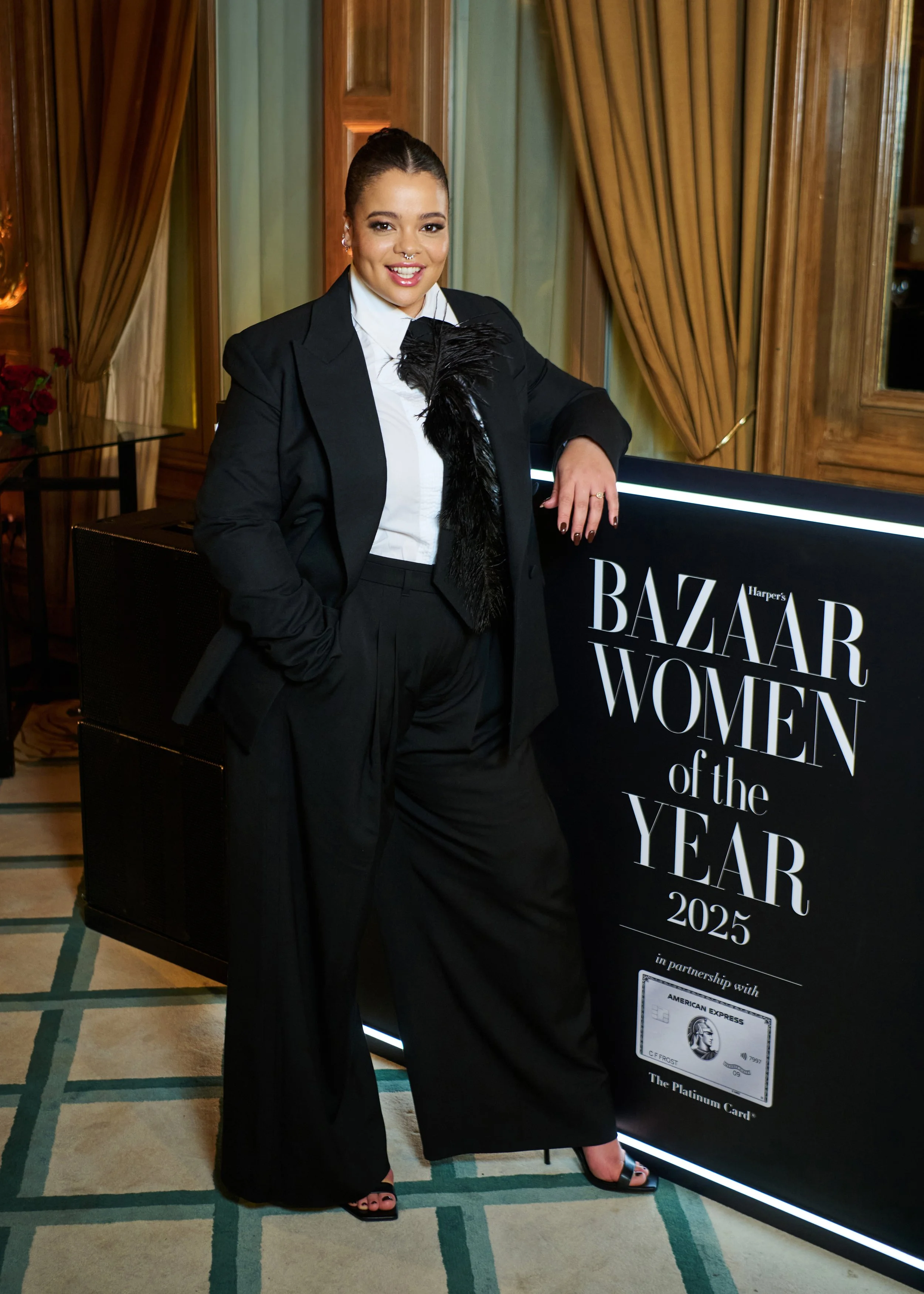 A woman dressed in a black suit with a white shirt and black feather accessory is standing next to a Harpers Bazaar Women of the Year 2025 sign.