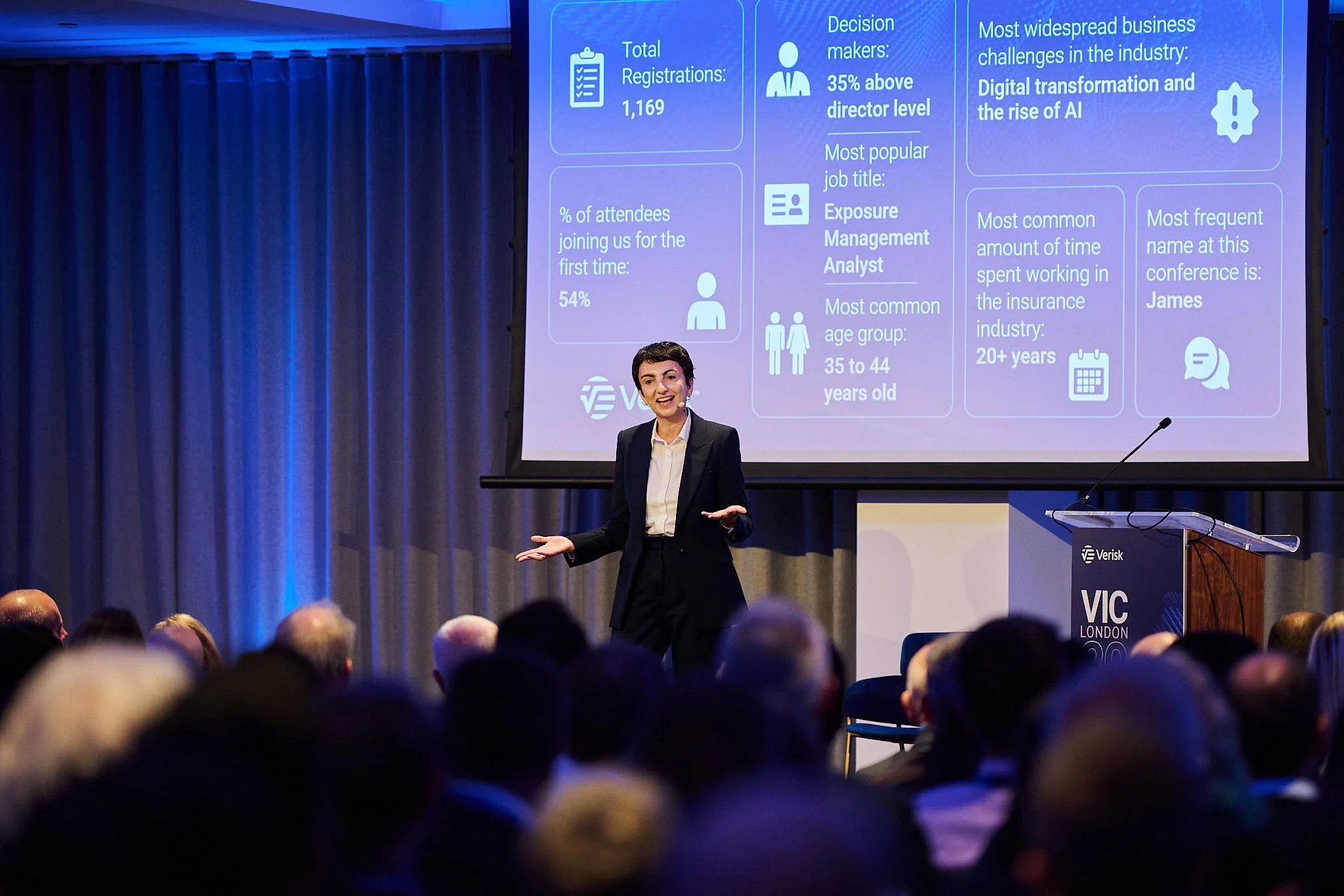 A woman in a black suit presenting at a conference in front of a large screen with data and statistics, with an audience seated in front of her.