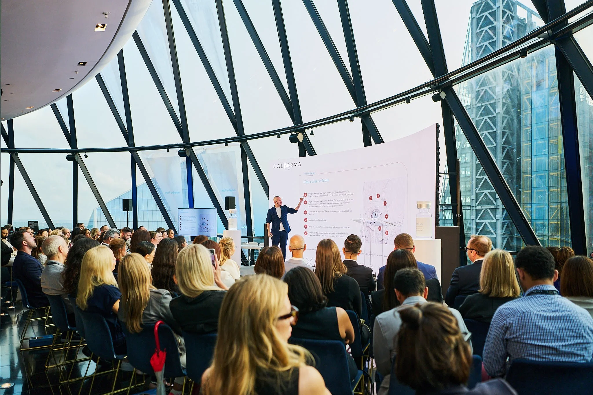 A speaker giving a presentation to a large audience in a modern, glass-walled conference room with a cityscape view.