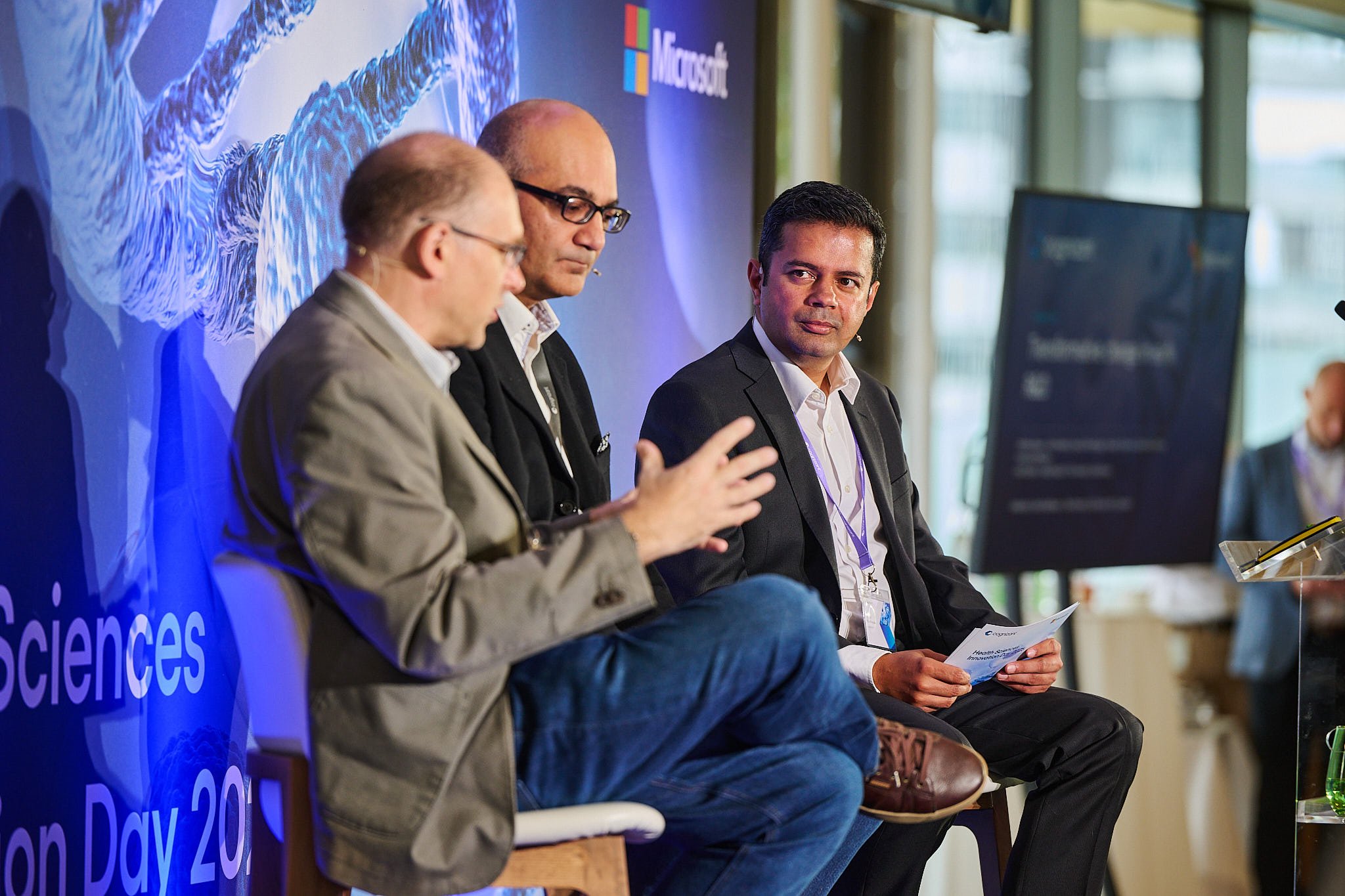 Three men seated on a stage during a conference, with a blue backdrop displaying the Microsoft logo and text about Science Day, engaged in discussion.