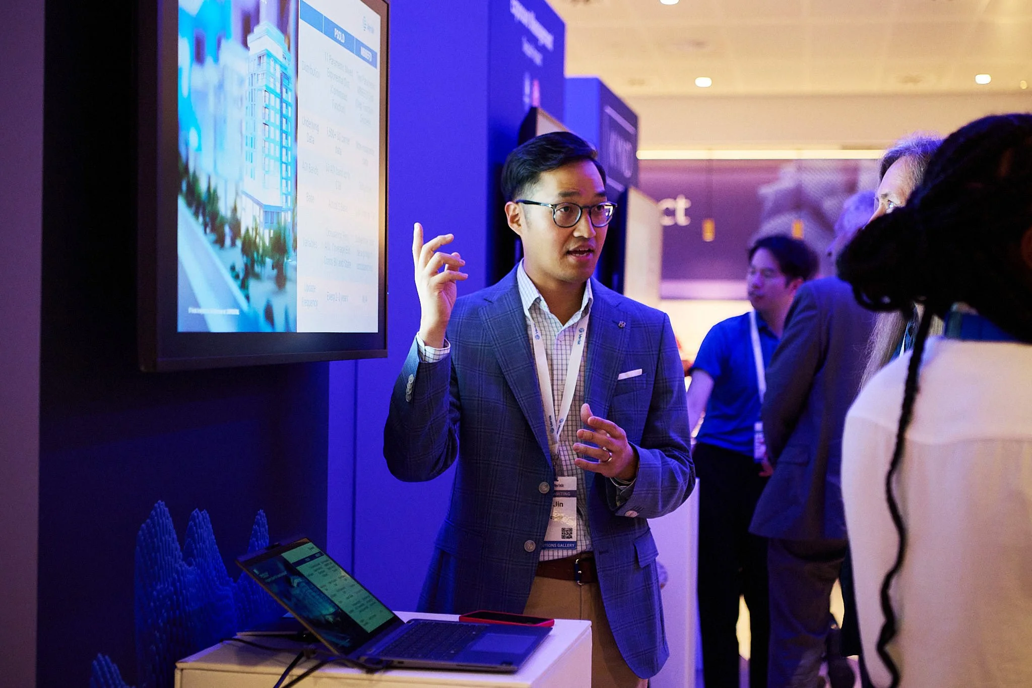 A man in a blue plaid blazer and glasses is speaking and gesturing with his hand at a business conference or trade show, standing next to a large screen displaying a cityscape and a laptop on a small table.