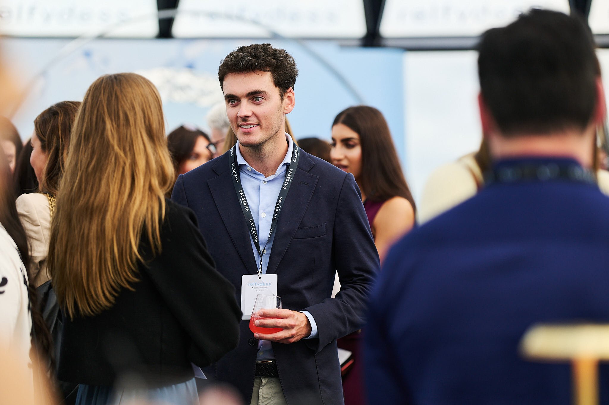 A young man in a navy blazer and light blue shirt holding a pink drink is talking to a woman with long red hair at a professional networking event with other people in the background.