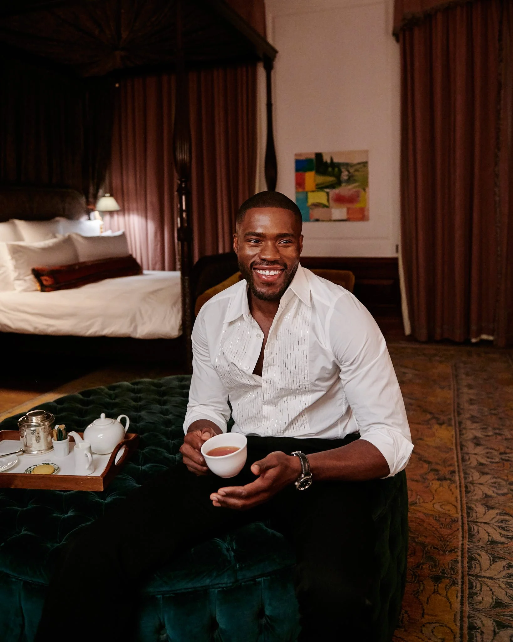 A man smiling and sitting on a green velvet ottoman in a hotel room, holding a teacup. There is a tray with teapot, sugar, and creamer on the ottoman. The background shows a bed with white linens and pillows, a painting on the wall, and curtains.