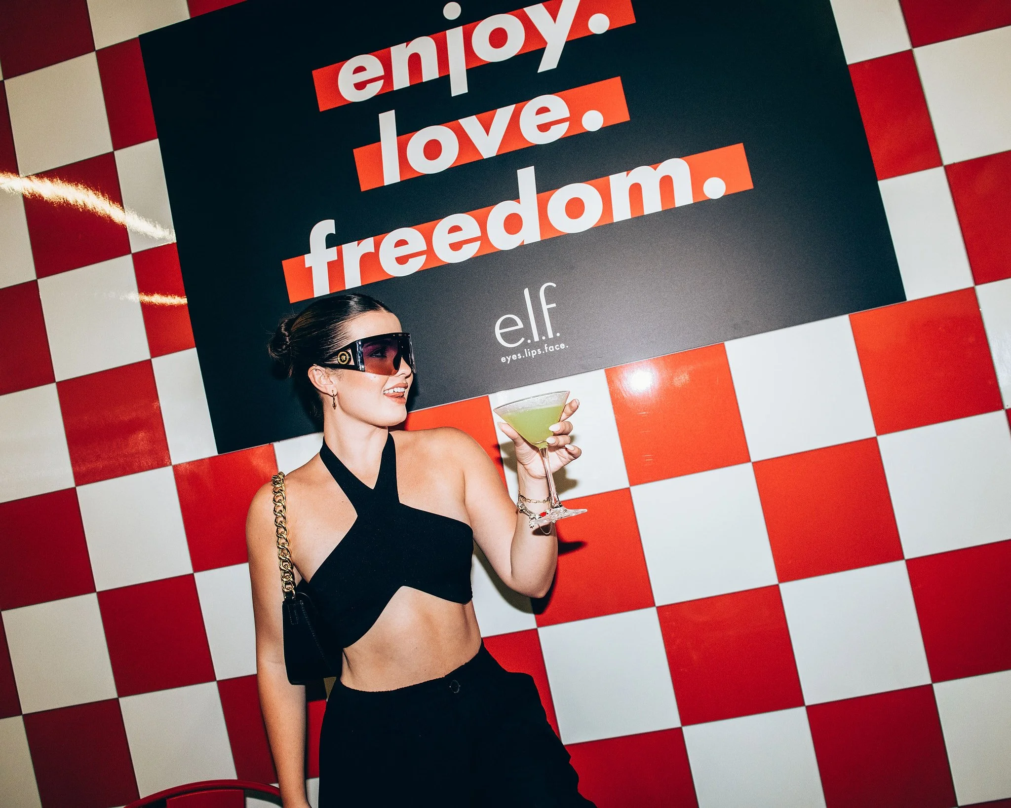 A woman dressed in a black halter top and pants, wearing large sunglasses, holds a green cocktail while smiling against a red and white checkered background with a sign reading 'enjoy. love. freedom.' and the 'e.l.f' logo.