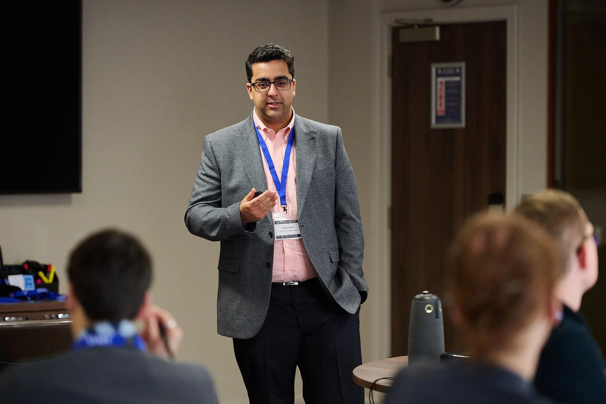 A man in a grey blazer and pink shirt giving a presentation to a group of people in a conference room.