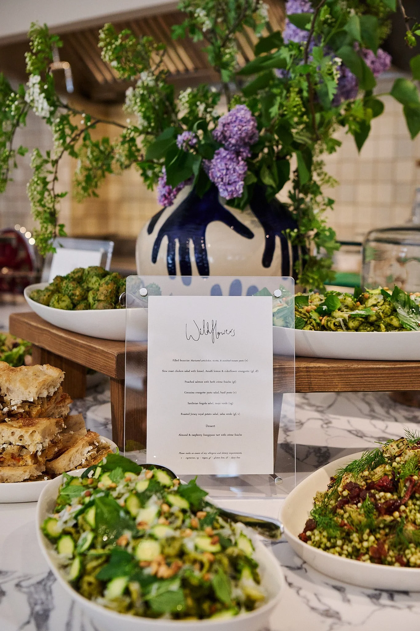 A display of various salads and dishes on a table, with a floral arrangement in a decorative vase in the background. There is a menu card indicating a menu named 'Wildflowers' with various food items listed.