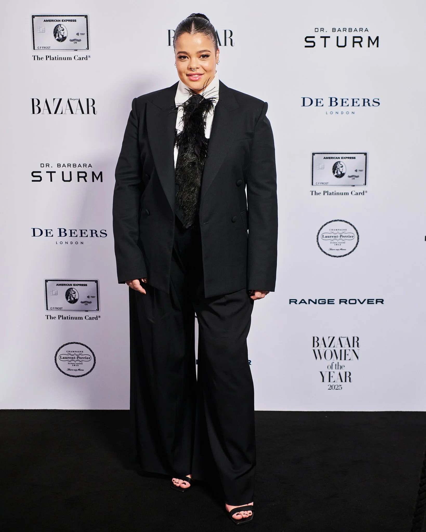 Woman in a black suit and wide-legged pants standing on a black carpet at the Harper's Bazaar Women of the Year 2025 event, with a white backdrop featuring logos and sponsors.
