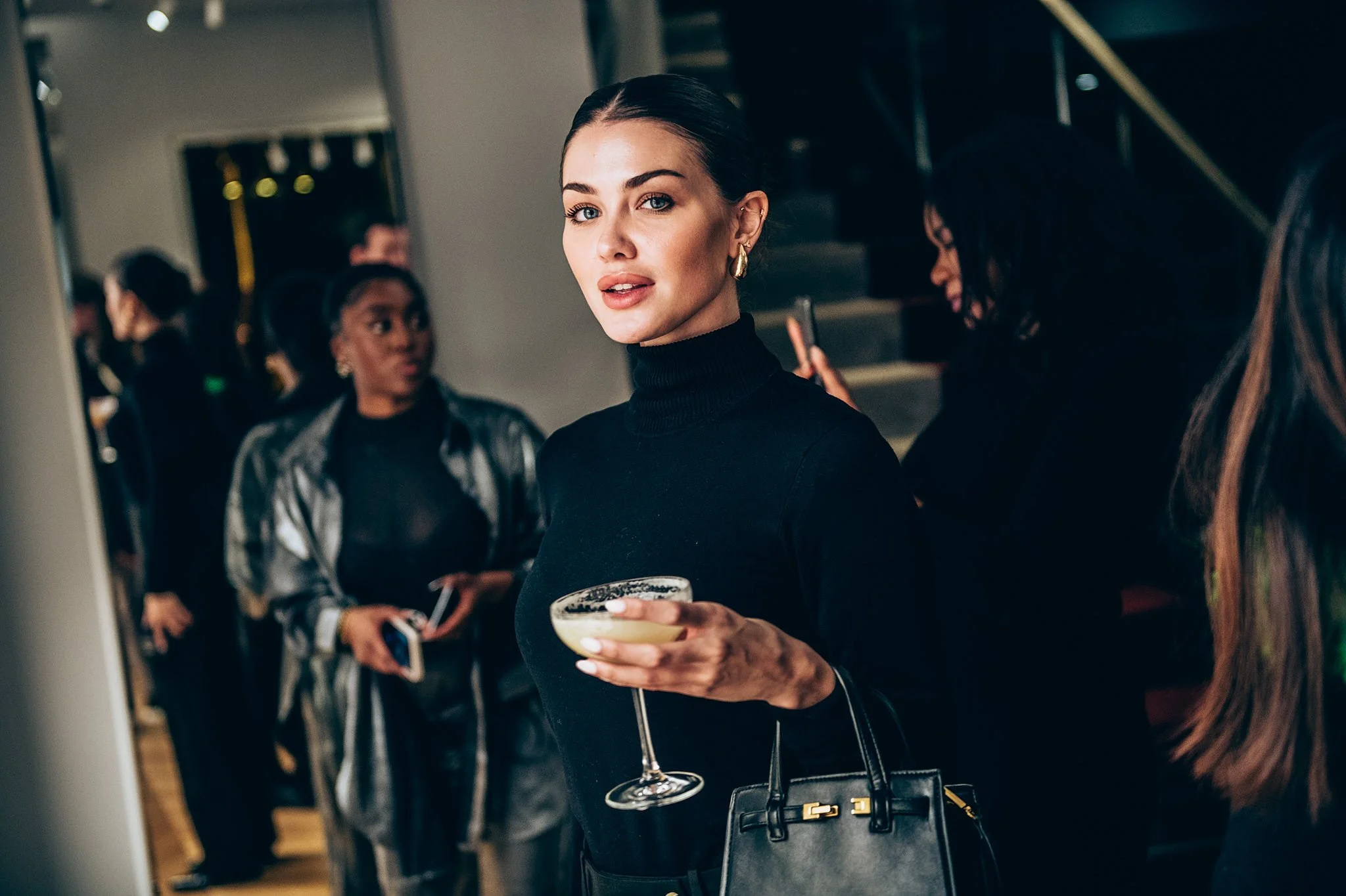 A woman with dark hair in a sleek bun, dressed in black, holding a martini glass, at a social gathering or party.