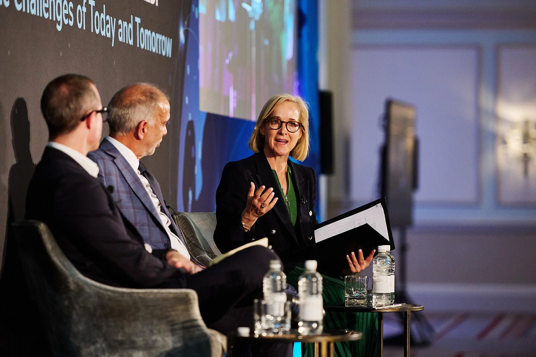 A panel of three people sitting on stage at a conference, with a woman speaking and two men listening. The woman has blonde hair, glasses, and is holding a folder. The men are wearing suits, and water bottles are on a table in front of them. There is