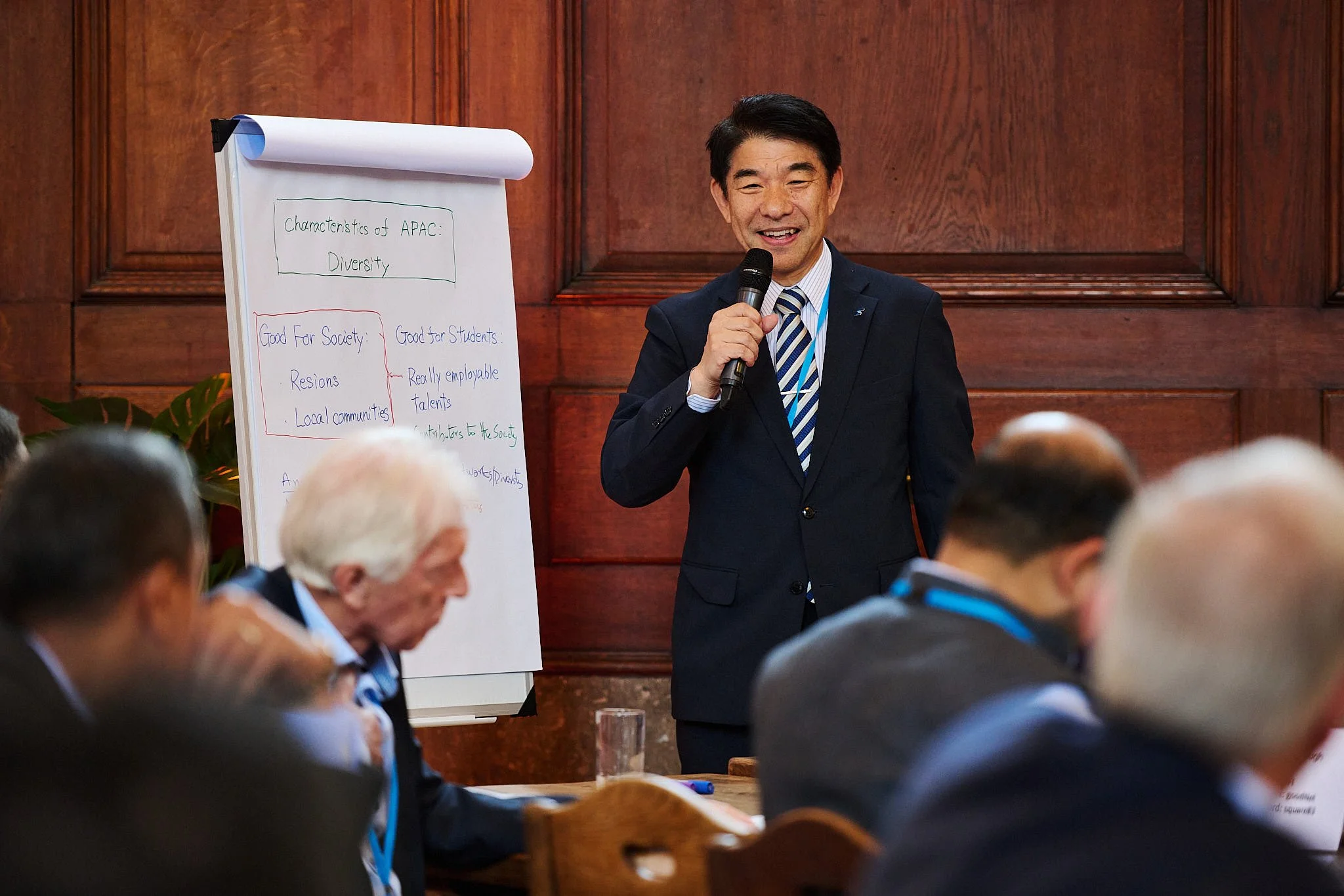 A man in a dark suit and striped tie is speaking into a microphone in a conference room with wood-paneled walls. Several people are seated in front of him, some with gray or white hair. A large whiteboard with handwritten notes on diversity and other