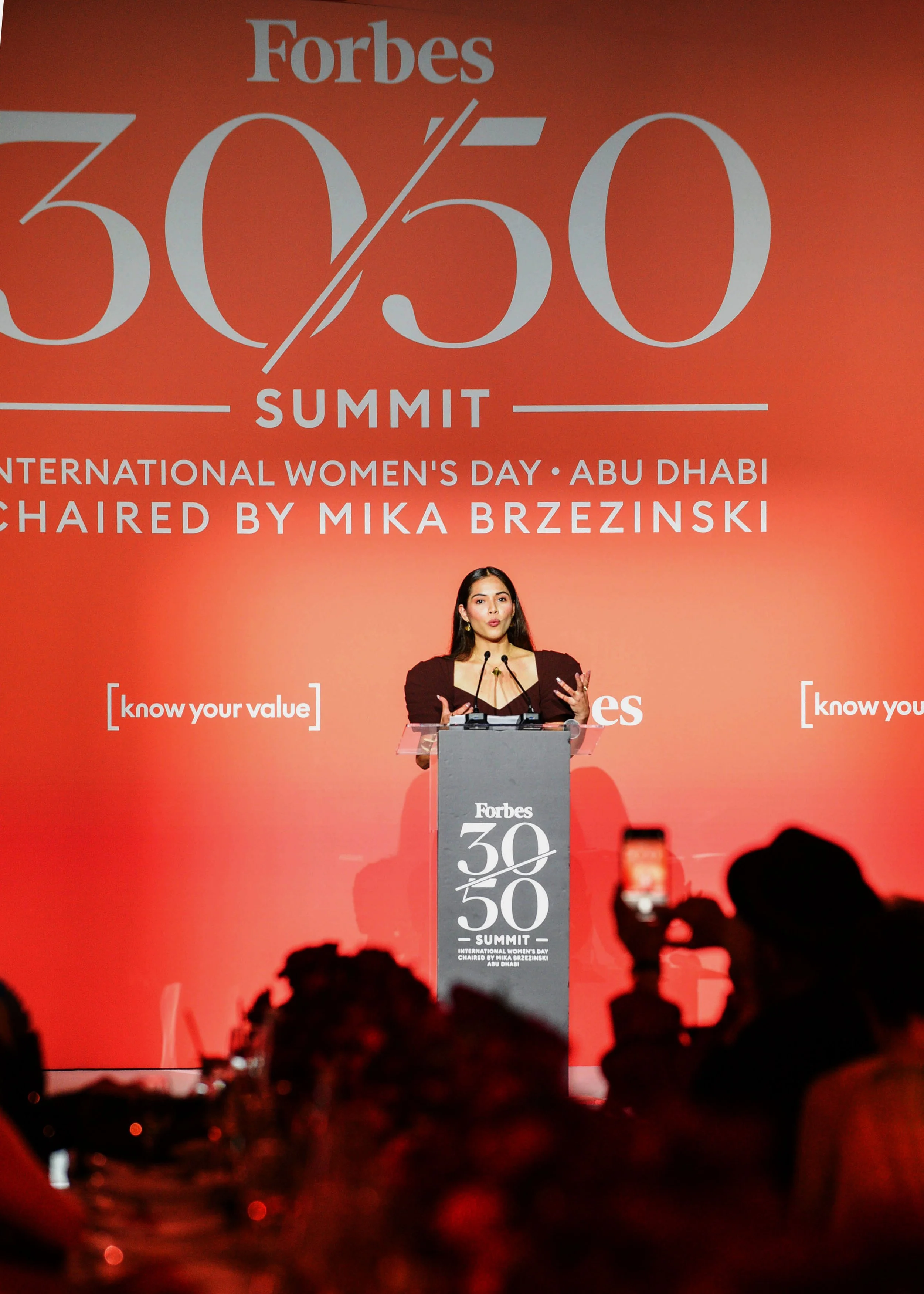 A woman speaks at a podium during a Forbes 30/50 Summit event in Abu Dhabi, celebrating International Women's Day. The backdrop is red with white text and the Forbes logo.