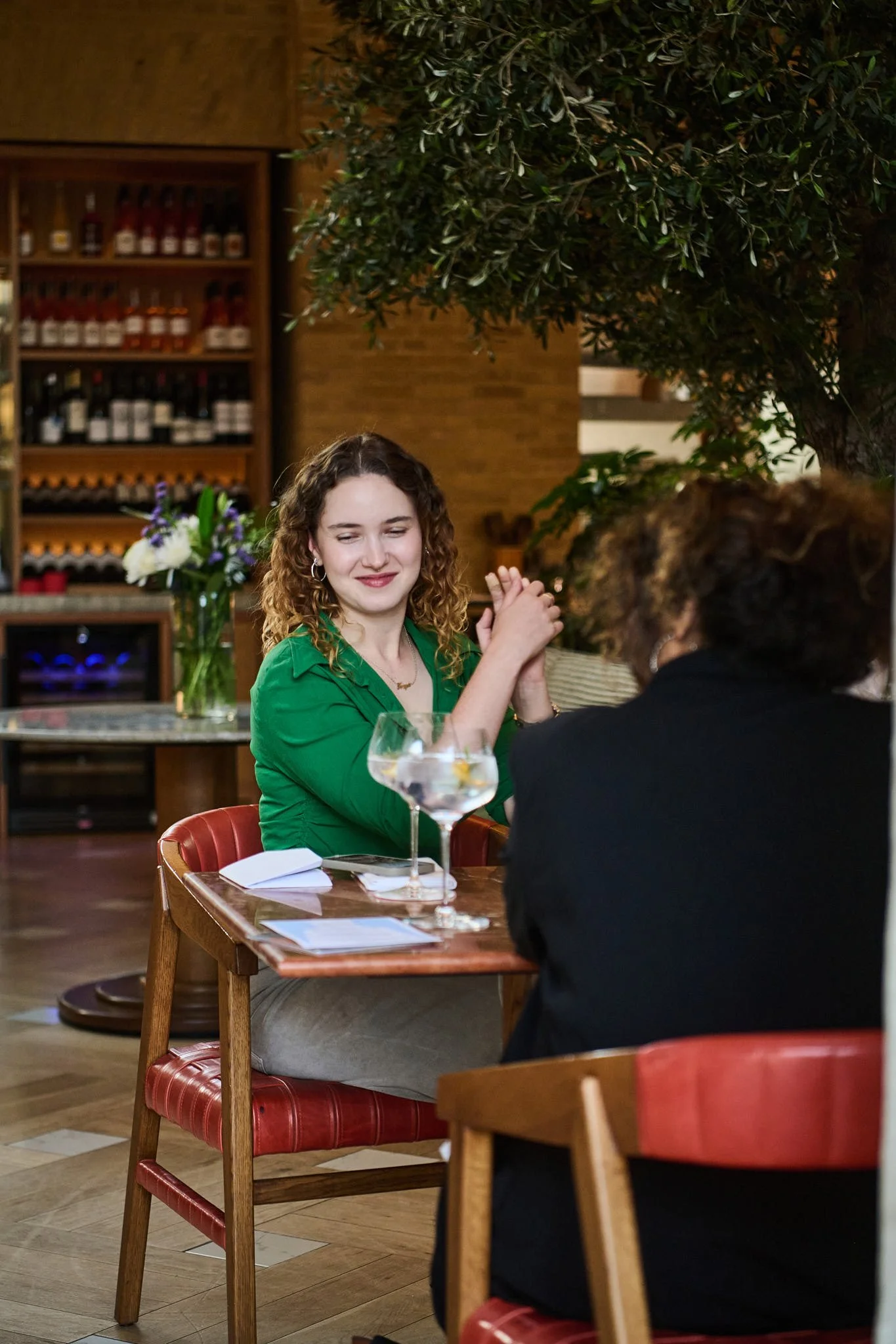 Two women sitting at a dining table in a restaurant, engaging in a friendly conversation. One woman is wearing a green blouse, and the other woman has curly hair. There are books and two glasses of water on the table, with a bouquet of flowers and a 
