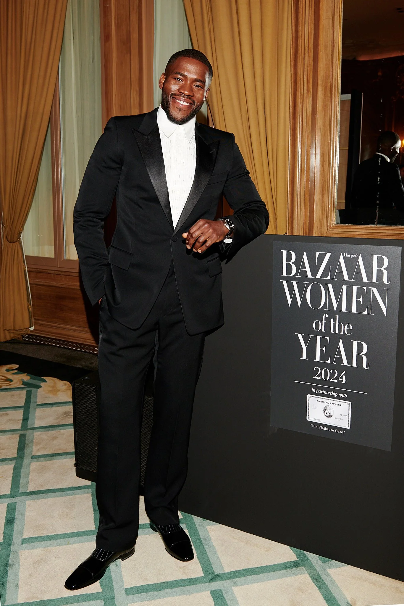 A man in a black tuxedo standing next to a sign that reads 'Bazaar Women of the Year 2024' at an event.