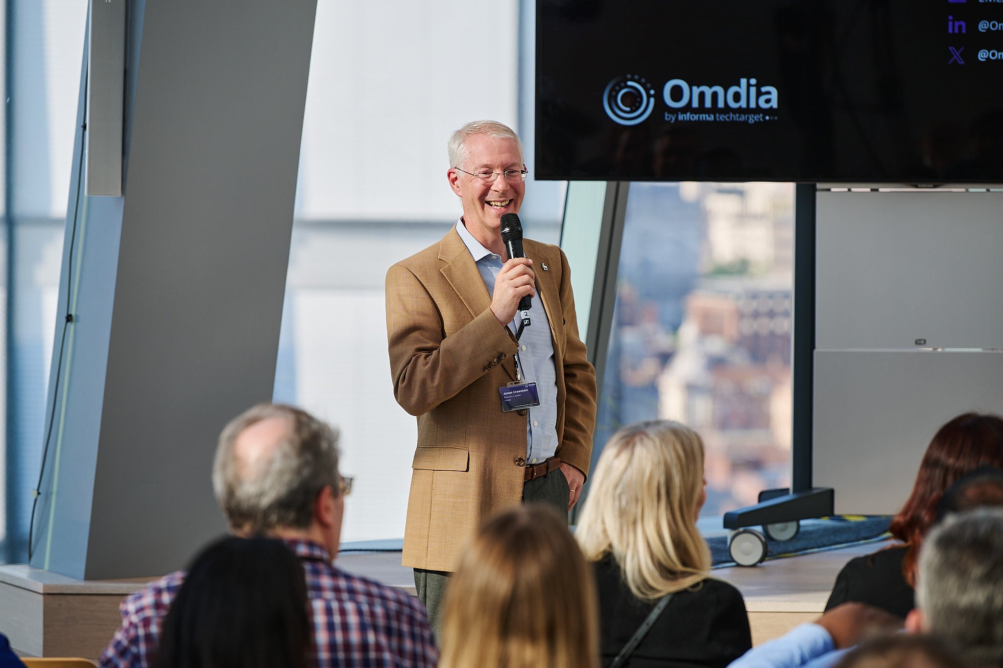 A man in a tan blazer and blue shirt holding a microphone, smiling during a presentation at a conference. Audience members are visible in the foreground, and a large screen displays a logo that reads 'Omdia by informa techtarget' in the background.