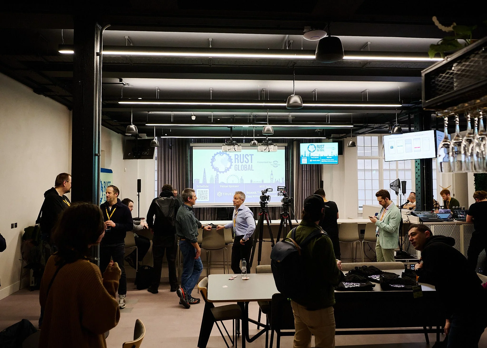 People gathering in a conference room with a presentation screen displaying 'RUST GLOBAL' and various digital screens, some individuals speaking, others using phones or cameras, and technical equipment on the side.