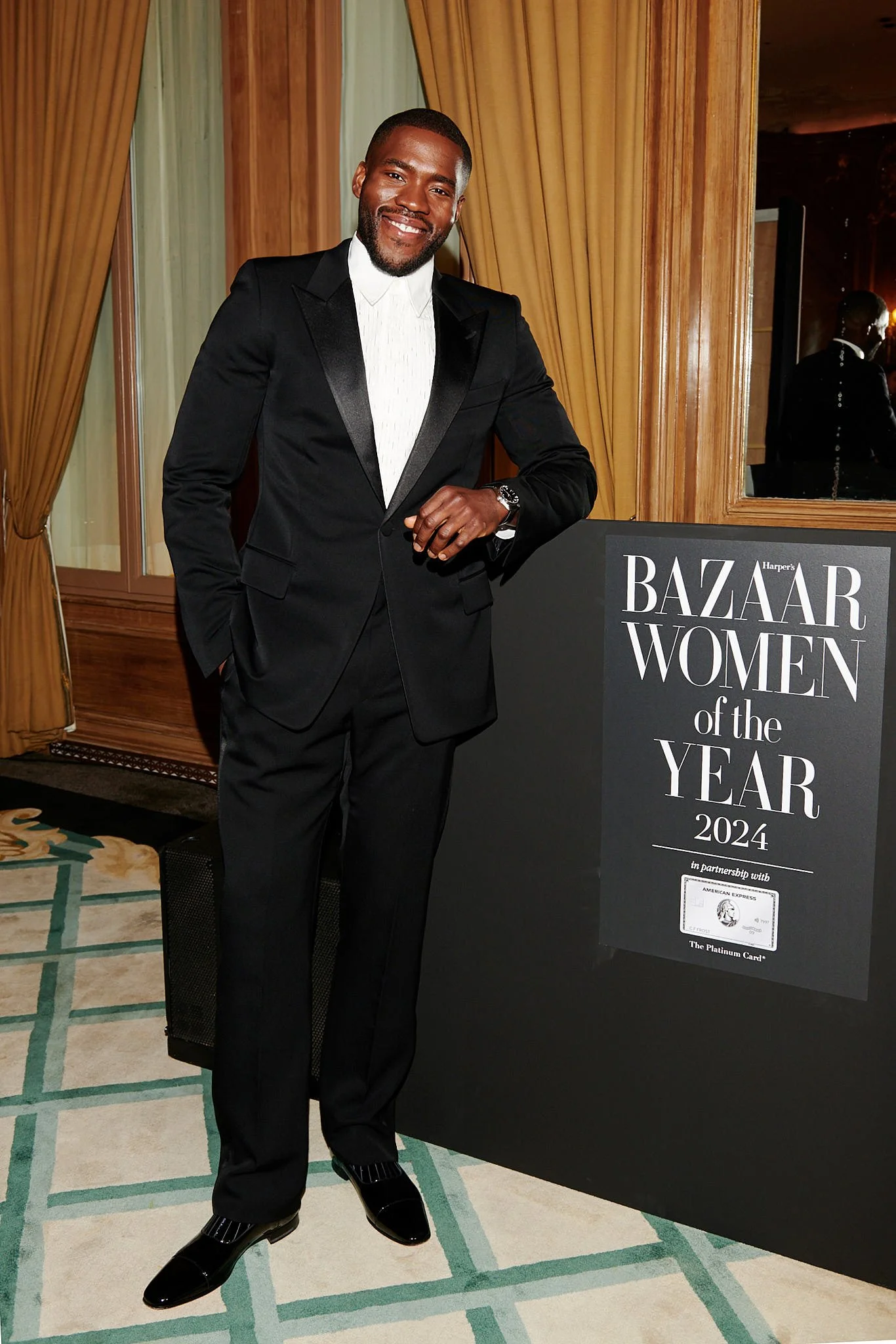 A man in a black tuxedo standing next to a sign that reads "Harper's Bazaar Women of the Year 2024".
