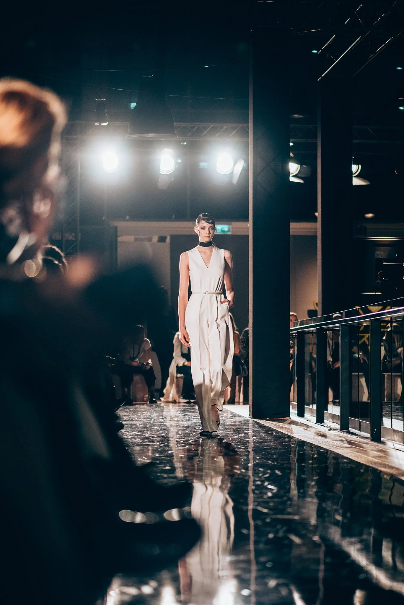 A female model walking down a runway during a fashion show, dressed in a white sleeveless jumpsuit and black choker, with bright stage lights overhead and audience members partially visible on the sides.
