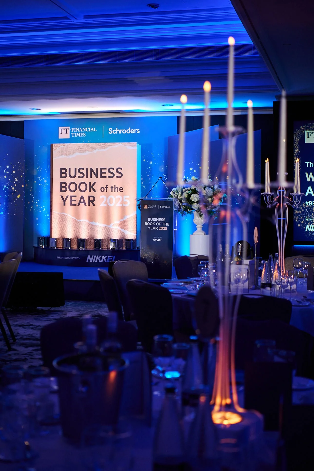 Conference room set for an event, with a large screen displaying 'Business Book of the Year 2025' and floral arrangements on tables, illuminated with blue lighting.