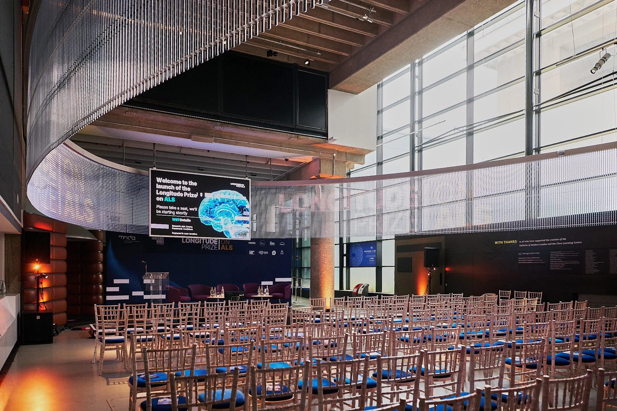 An empty conference room set up with rows of white chairs with blue cushions, facing a small stage with a podium and a screen displaying information about the Longitude Prize on ALS. The room has large floor-to-ceiling windows letting in natural ligh