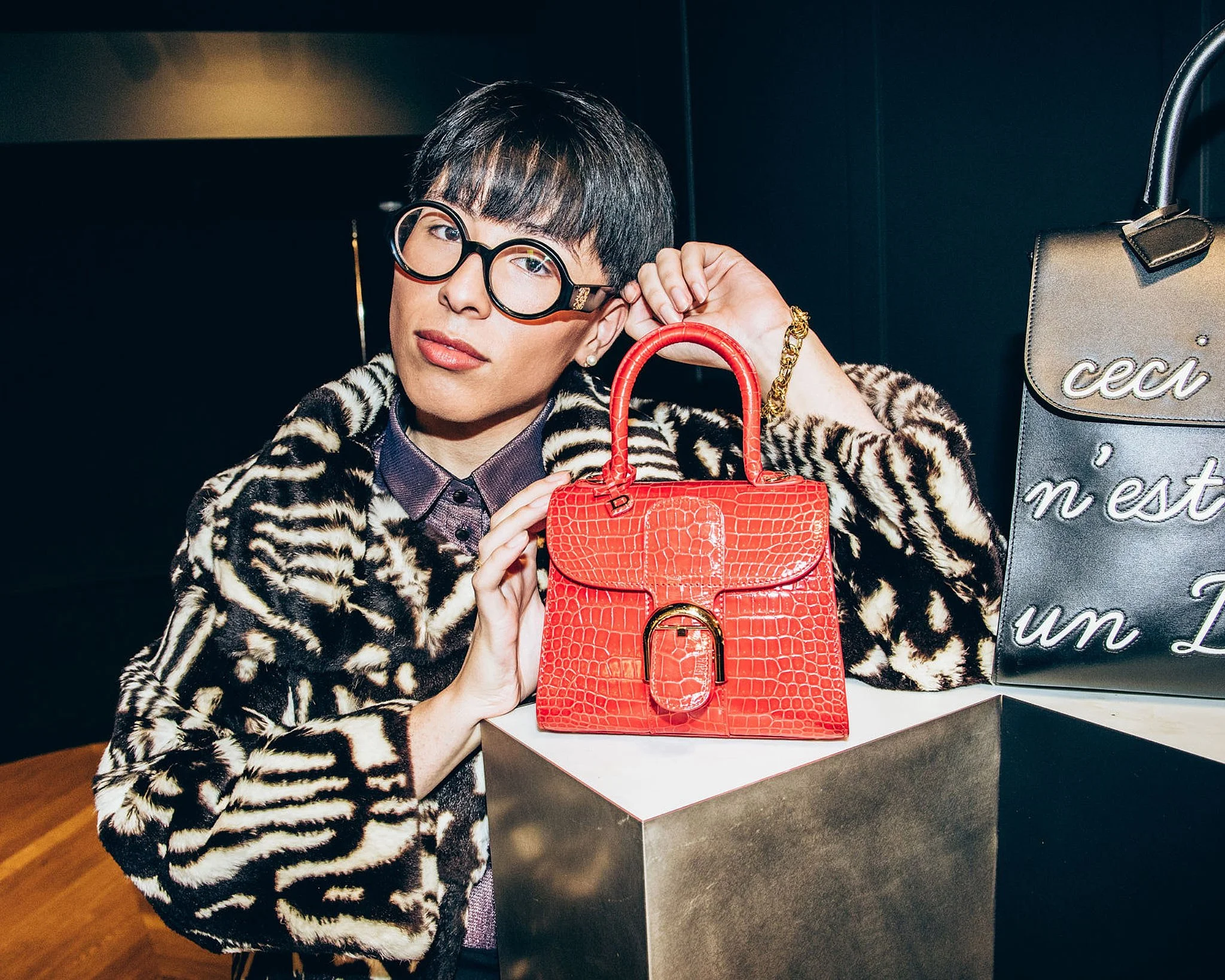 Person with black hair and glasses holding a red crocodile-patterned handbag, wearing a leopard-print coat, sitting at a display with other bags.