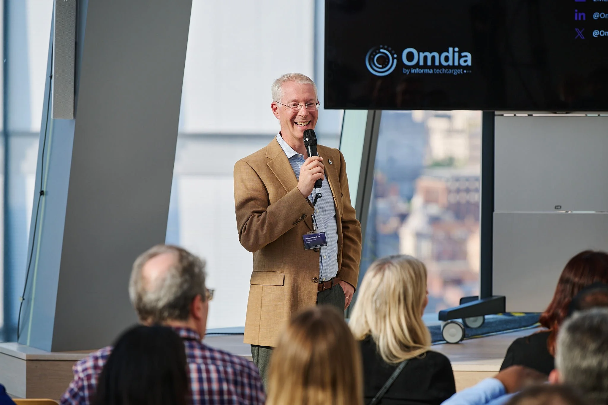 A man in a tan jacket and glasses smiling while holding a microphone, speaking at a conference with a screen displaying 'Omdia' in the background, and an audience listening.